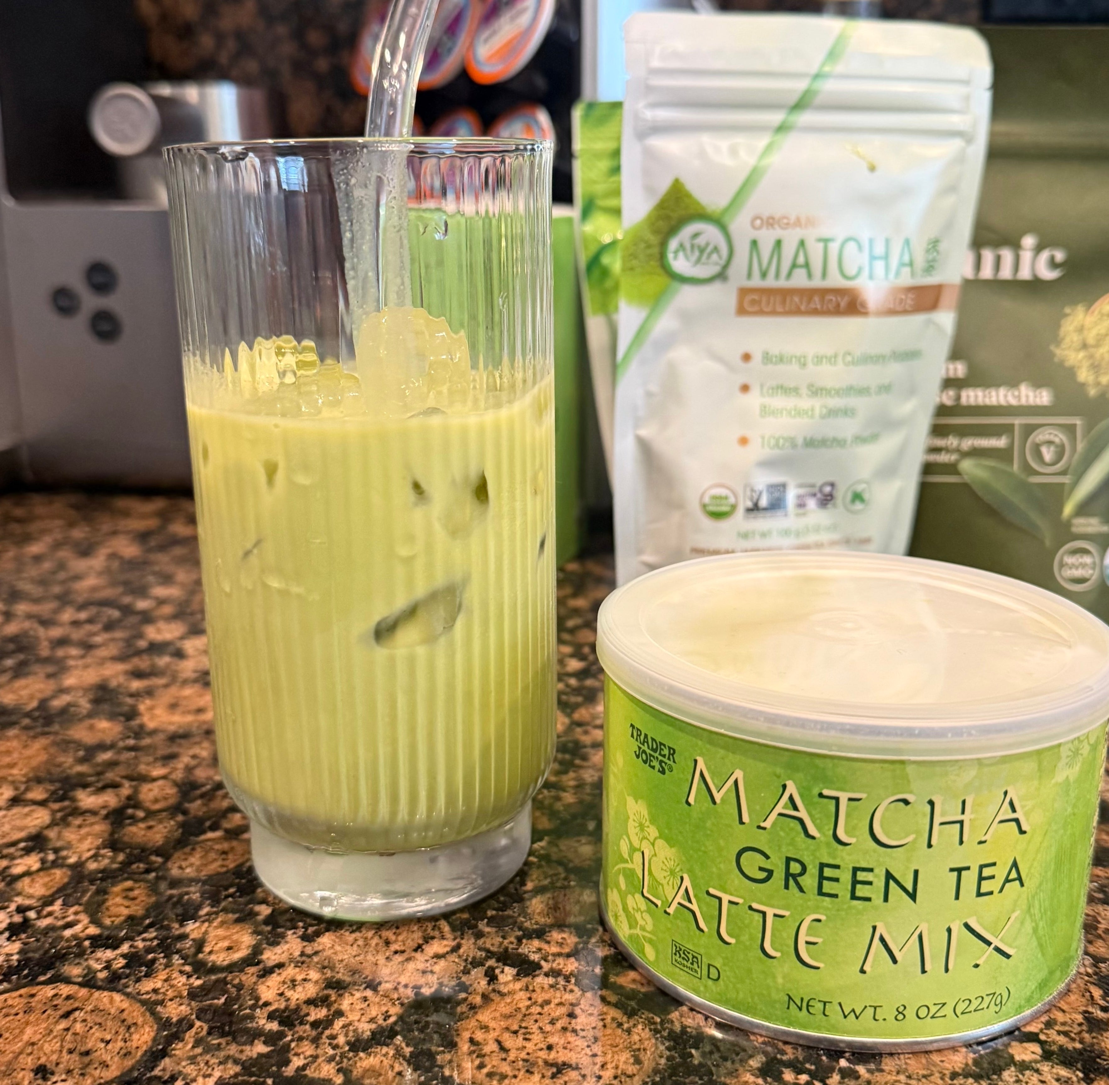 Glass of iced matcha latte on a countertop with matcha latte mix and ingredients in the background