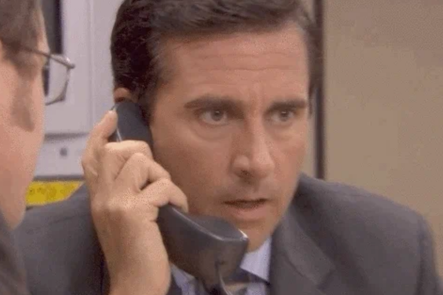 Man on phone, appearing surprised, in an office setting; scene from a comedic TV show