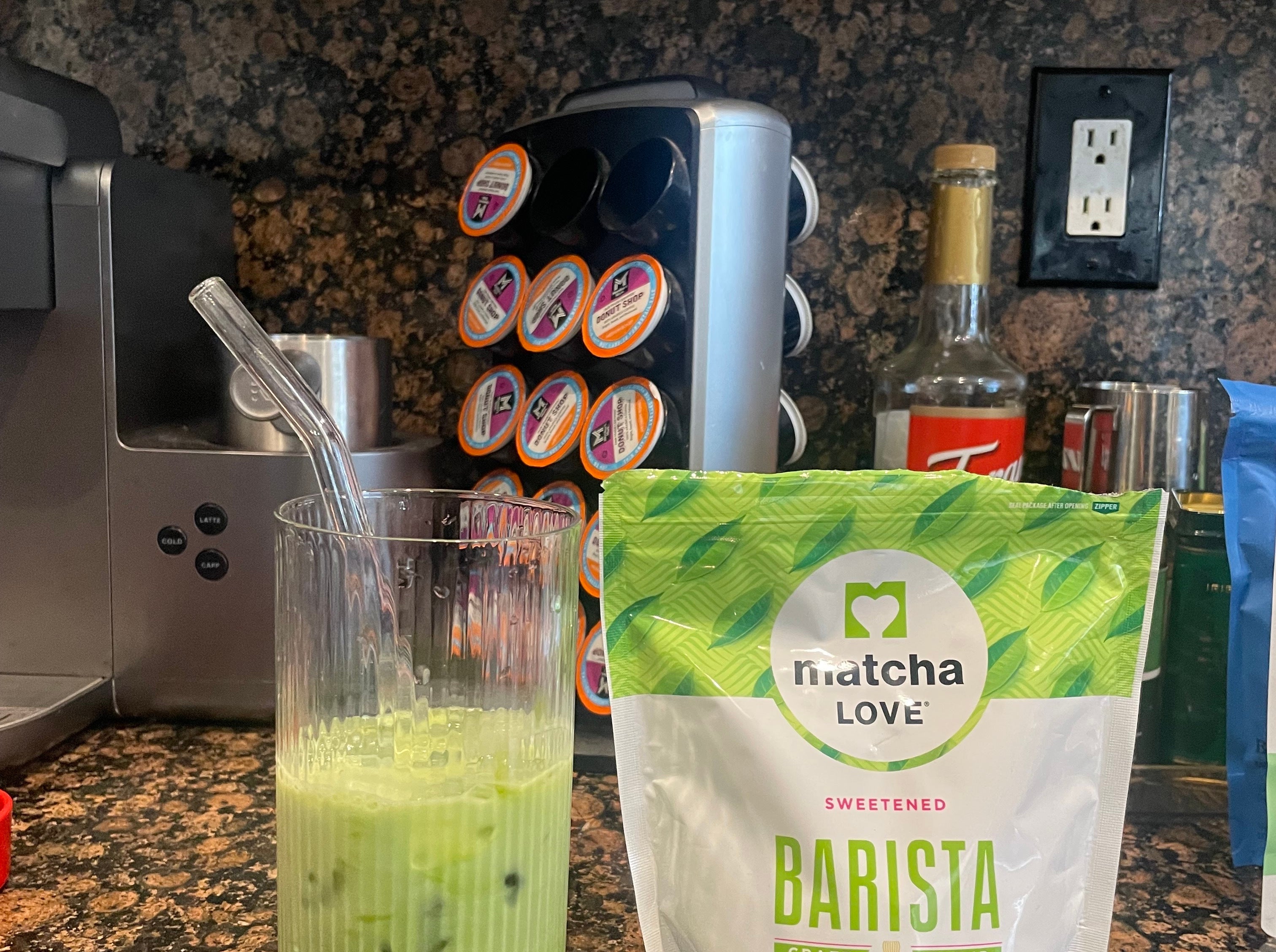 Iced matcha latte in a glass with a straw on a kitchen counter, next to a bag of Matcha Love powdered tea