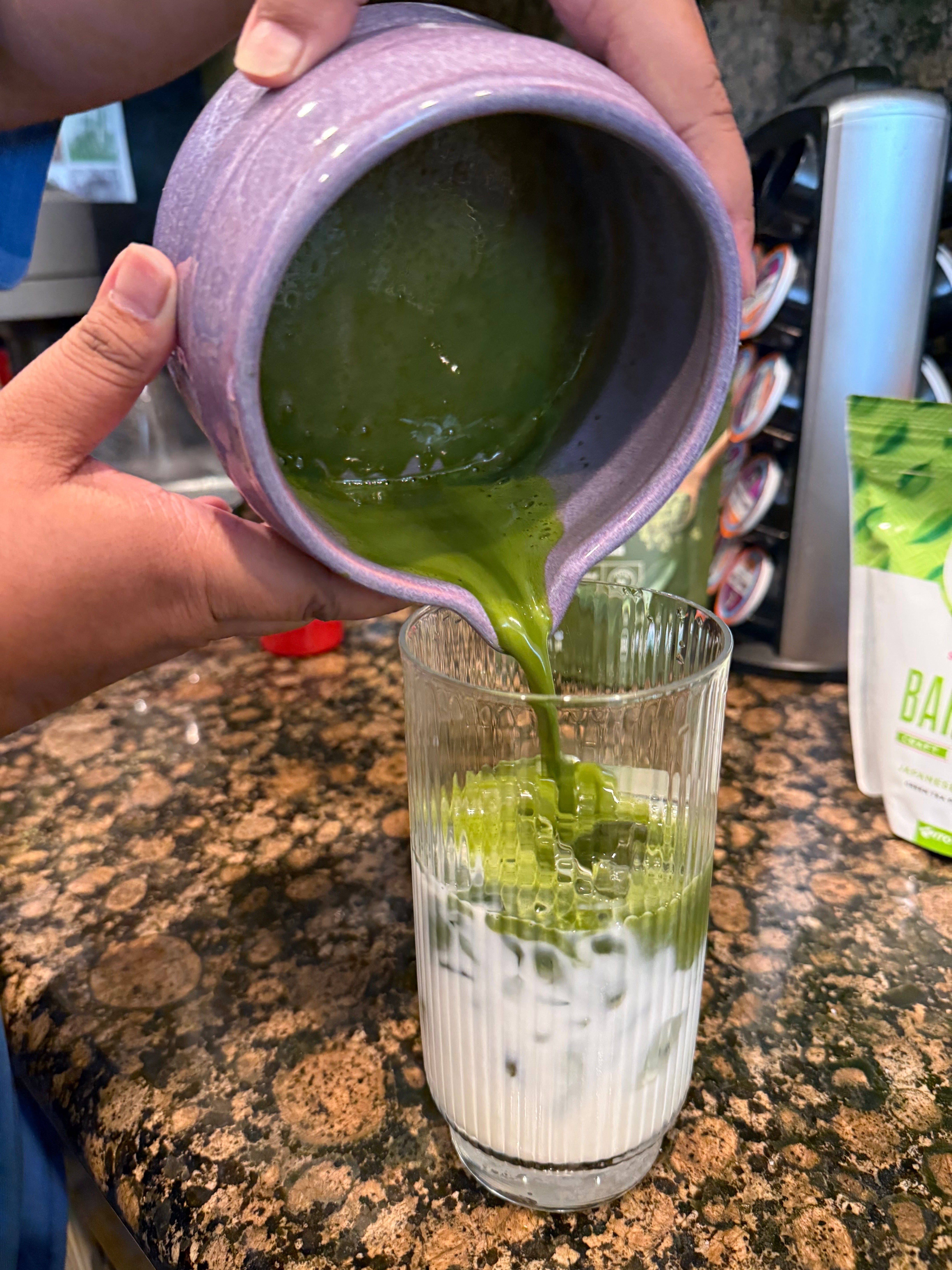 Matcha latte being poured into a glass of milk with ice on a kitchen countertop