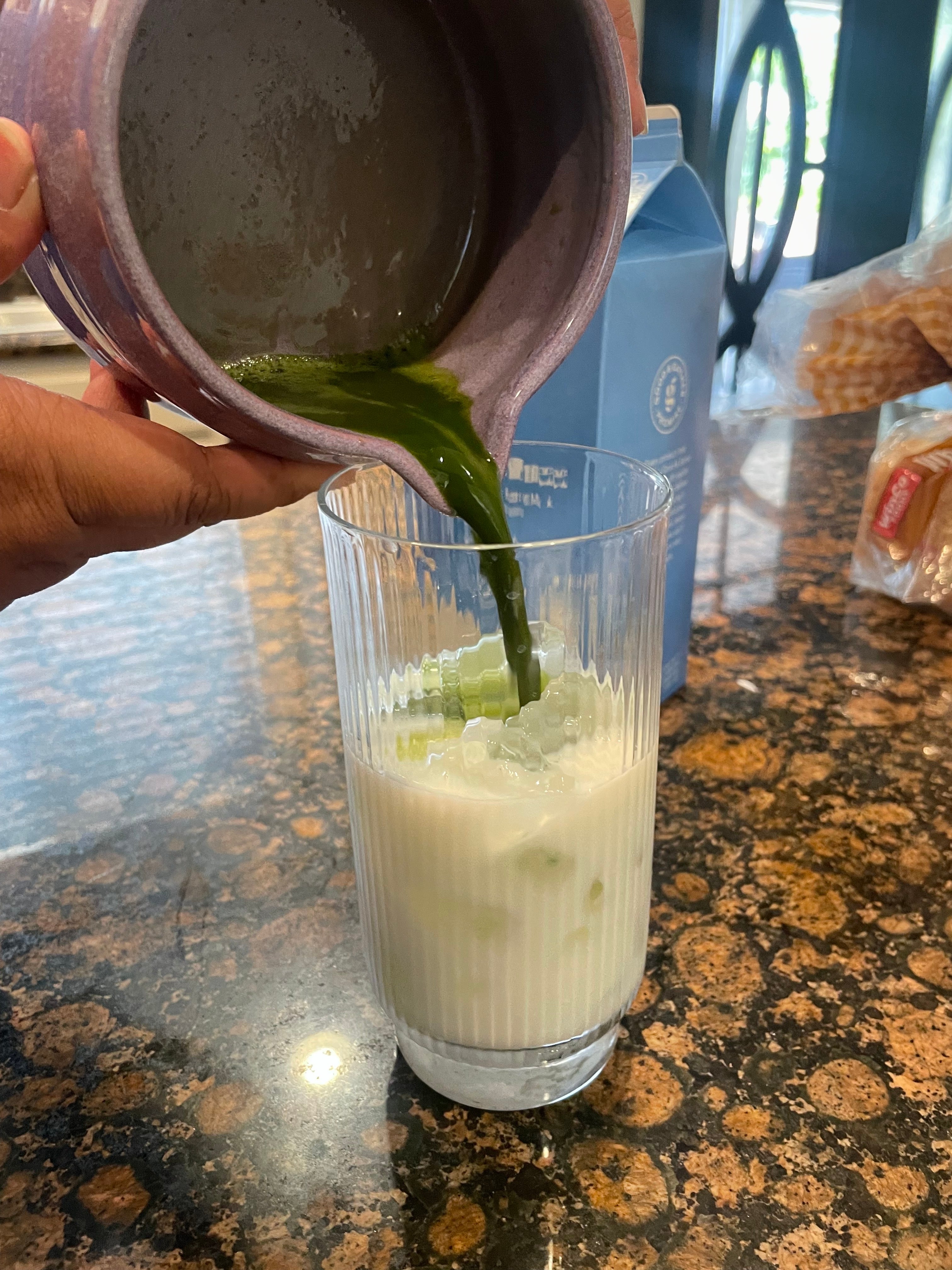 Matcha being poured into a glass of iced milk on a kitchen counter with food items in the background