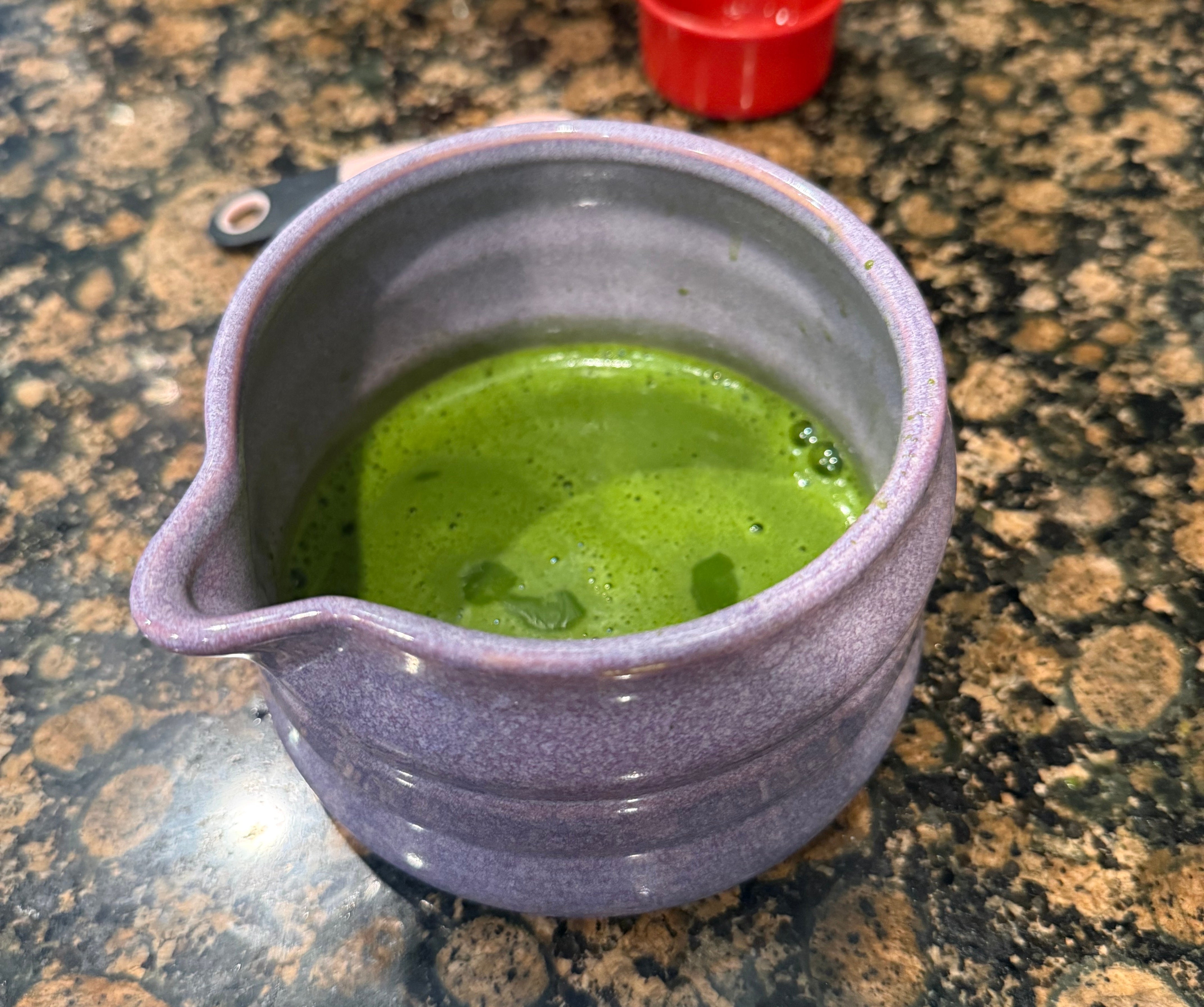 A ceramic bowl filled with frothy matcha tea sits on a speckled countertop, next to measuring cups and a coffee machine