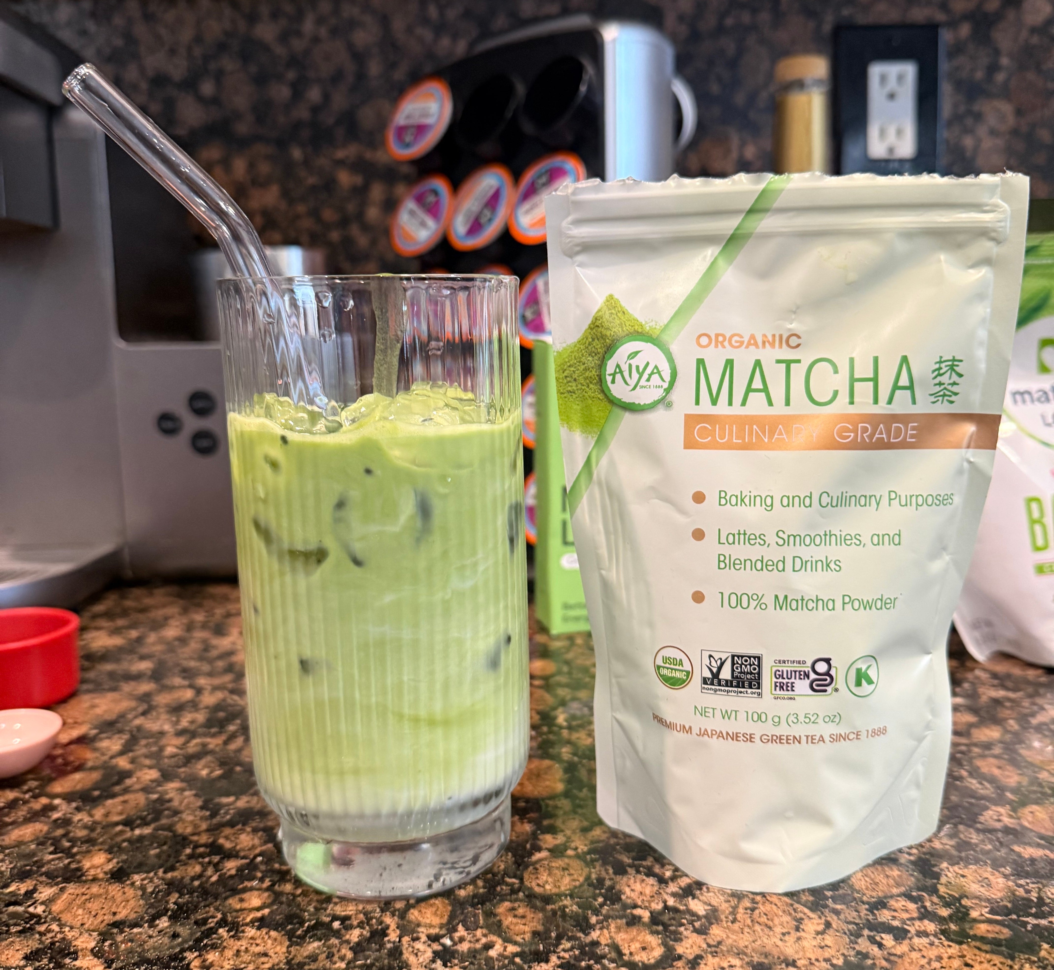 Iced matcha latte in a glass with a metal straw next to a bag of organic culinary-grade matcha powder on a kitchen countertop