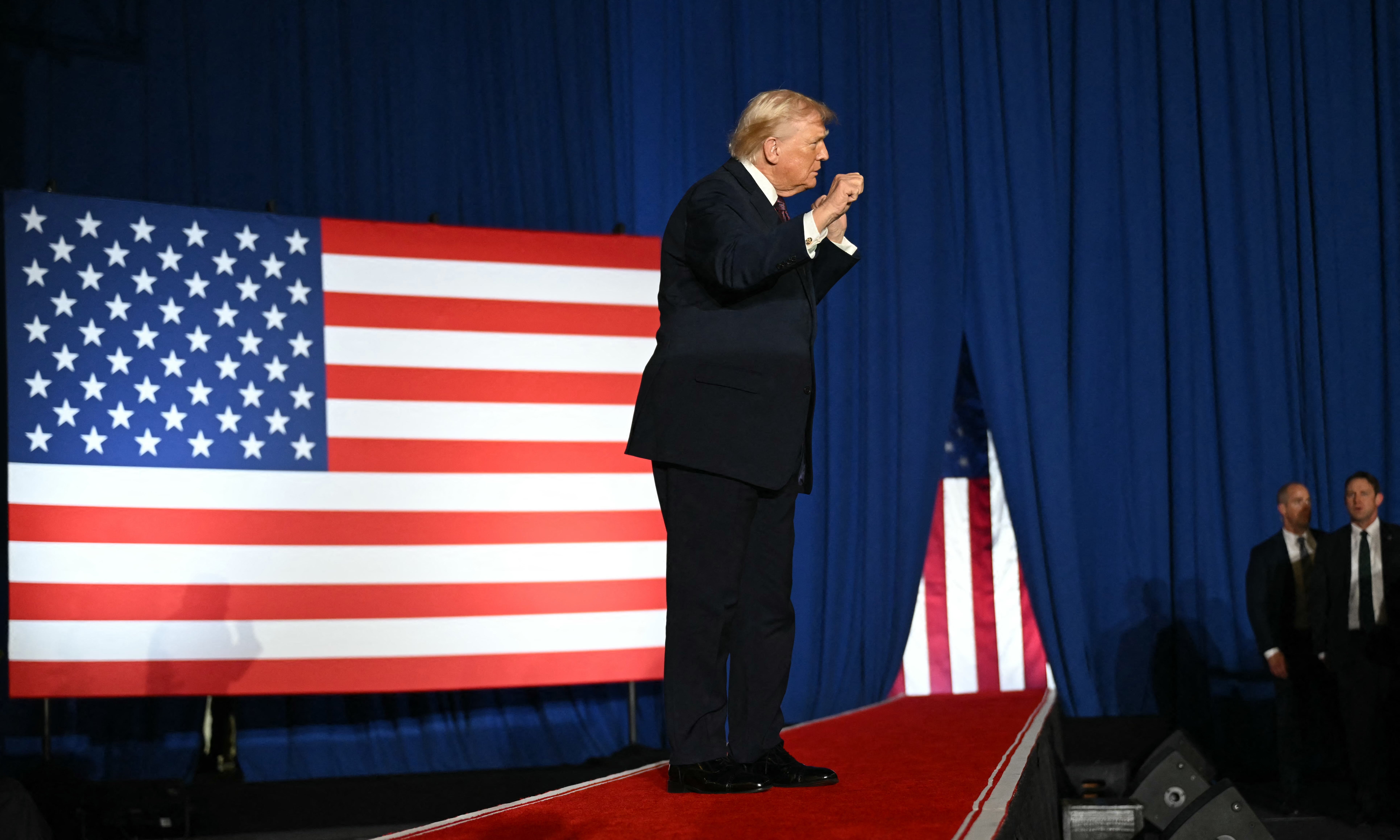 Person i dress som snakker energisk på scenen med et stort amerikansk flagg i bakgrunnen. Publikum og trygghet er tilstede