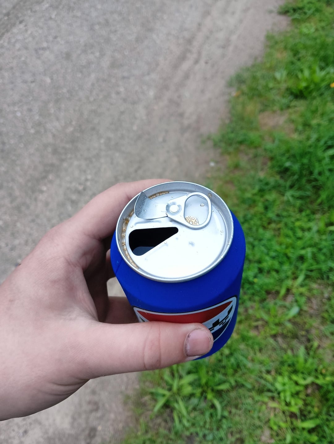 Tangan memegang kaleng soda terbuka dengan penutup biru, di luar ruangan di jalan berumput