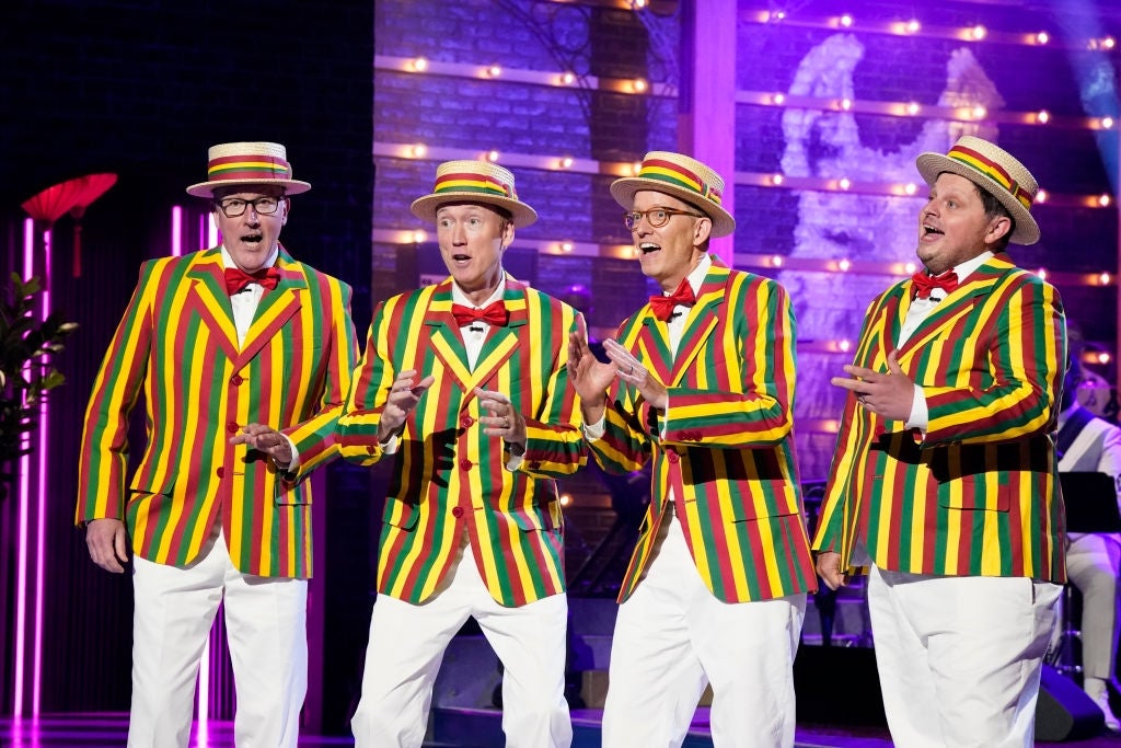 Group of four people singing in colorful striped blazers, bow ties, and boater hats on stage with a performer in the background