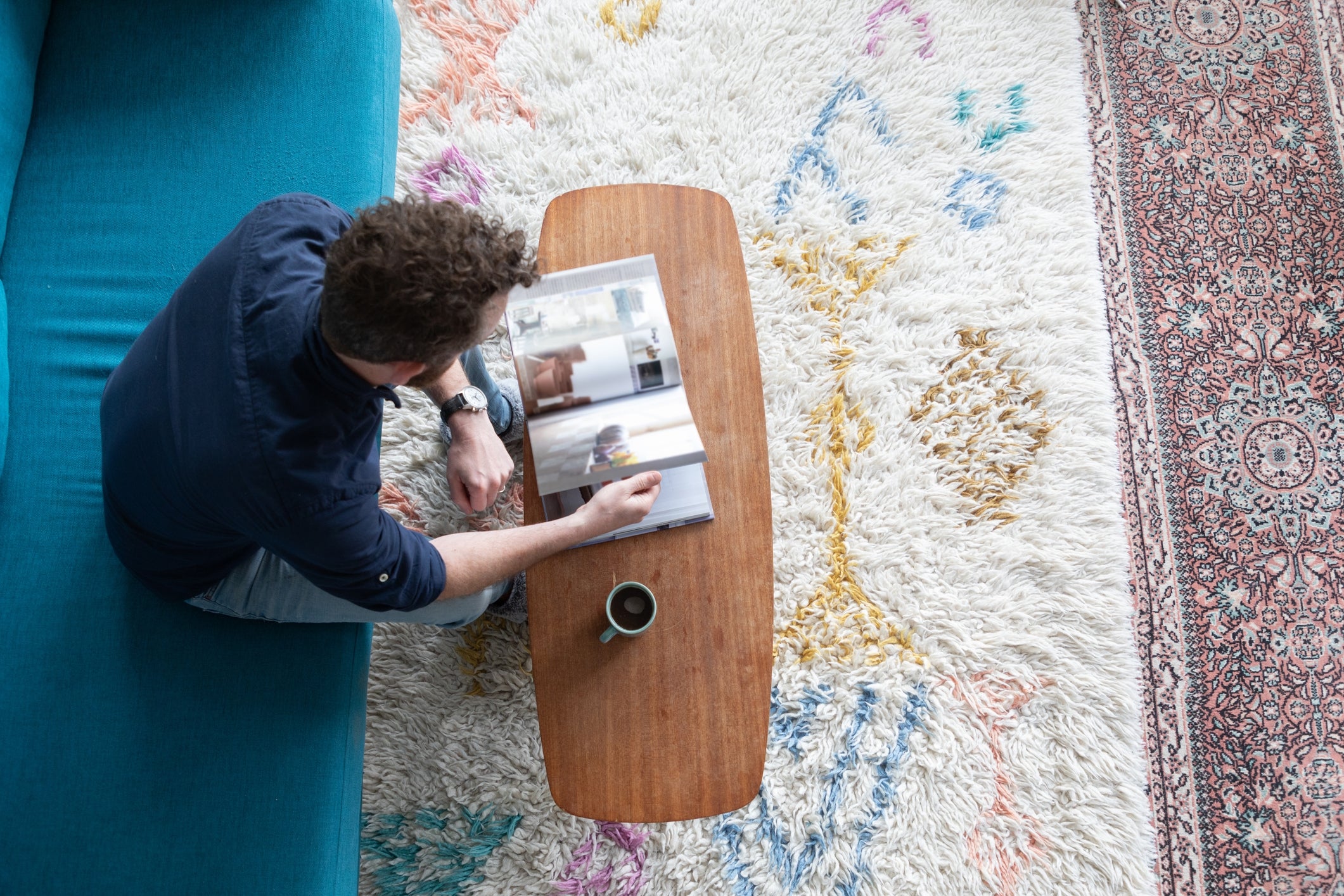 Person reading a magazine on a wooden table, next to a coffee cup, sitting on a couch with a patterned rug underneath