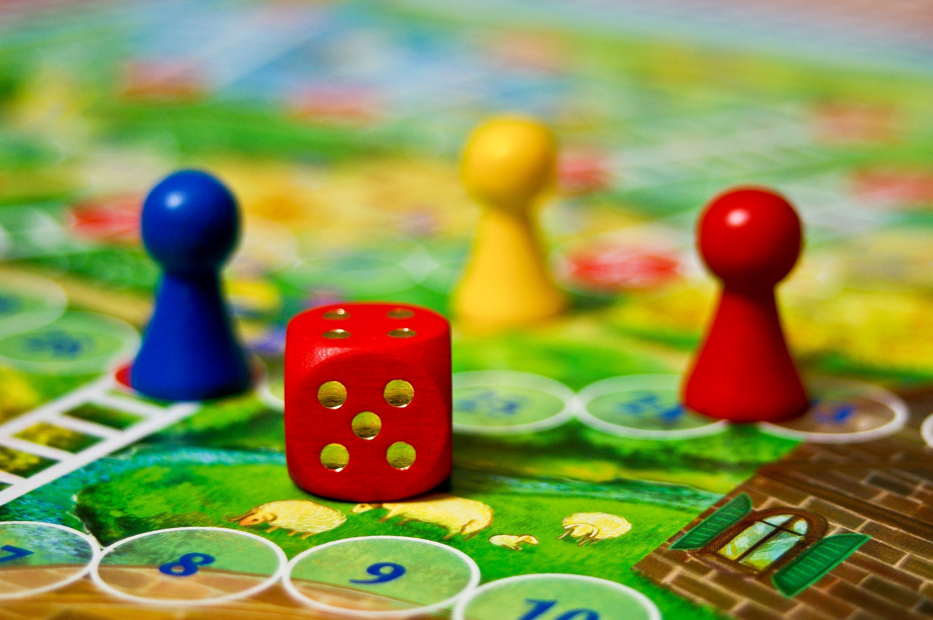Game board with colorful pieces, including red and yellow pawns and a red die showing six dots. The board features a path and numbered spaces