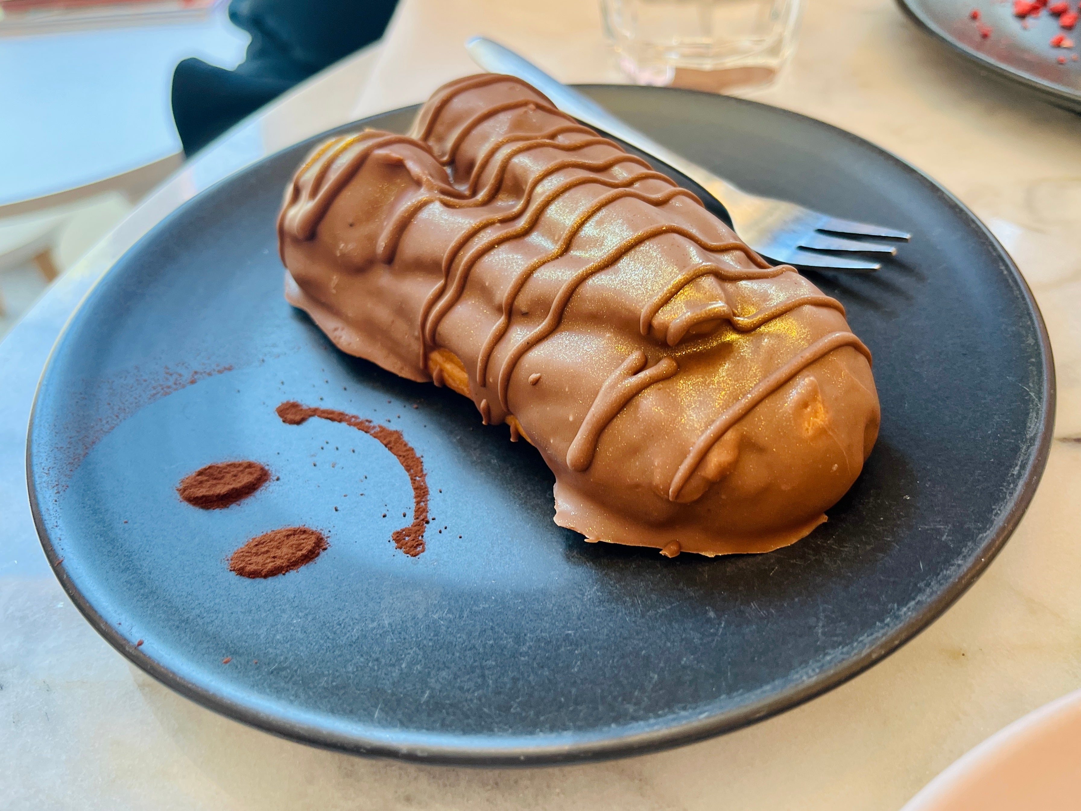Chocolate-covered éclair connected  a sheet  with cocoa pulverization  smiley face, fork beside it