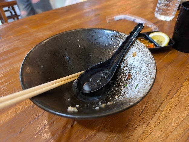 Empty ramen vessel  with chopsticks and a spoon connected  a woody  table, beside a tiny  crockery  with citrus  slices and condiments