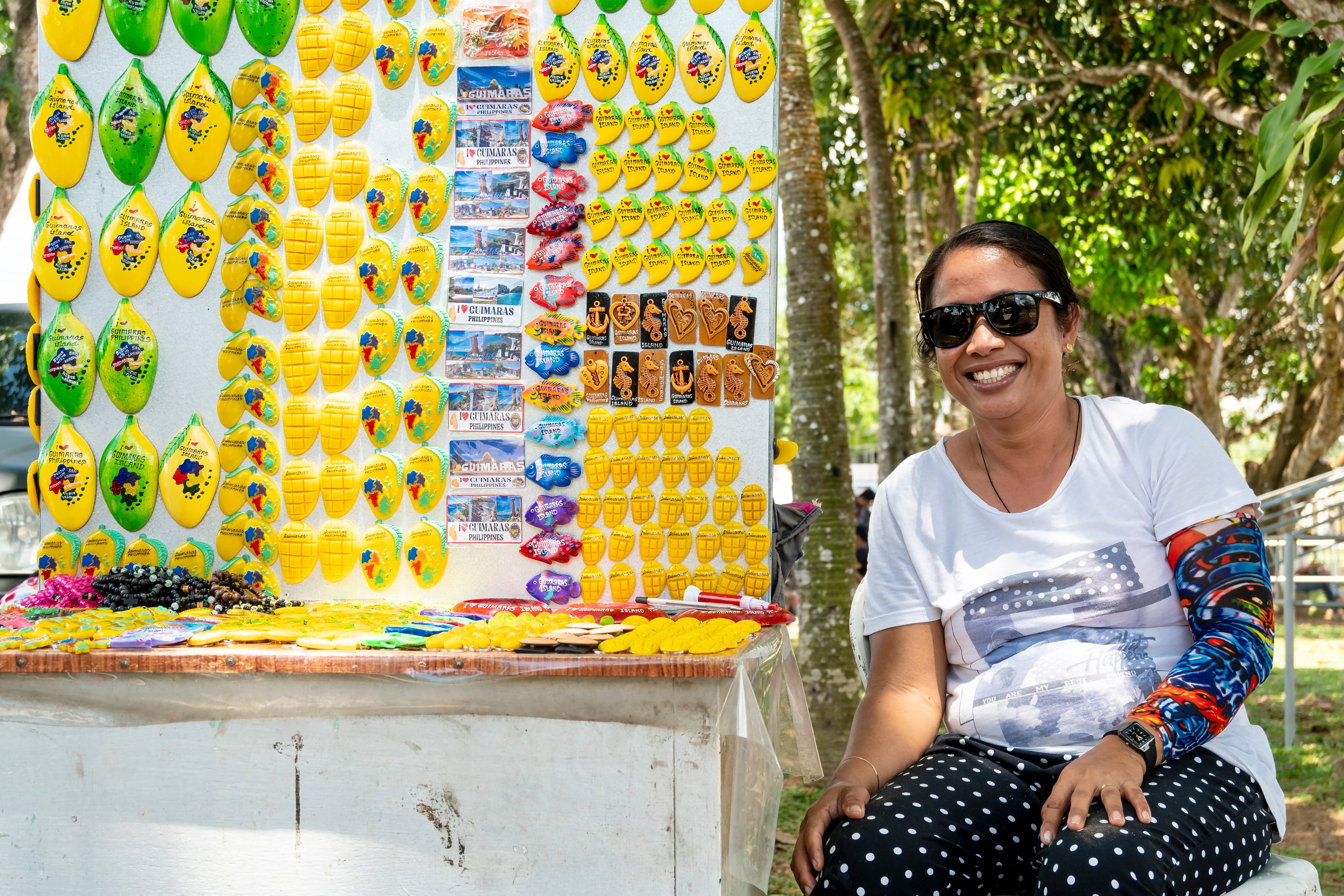 Smiling pistillate   with sunglasses sitting beside a basal   displaying colorful keychains and souvenirs successful  a sunny outdoor setting