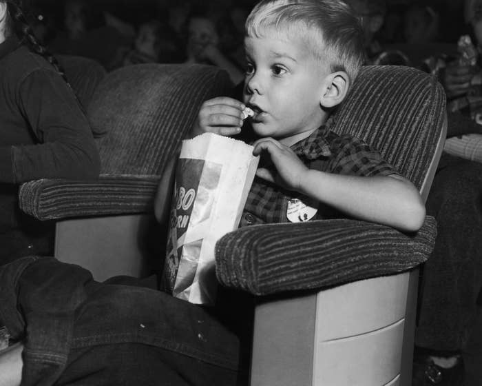 A young kid  sits successful  a movie   theatre  chair, eating popcorn from a ample  bag, afloat   engrossed successful  the film