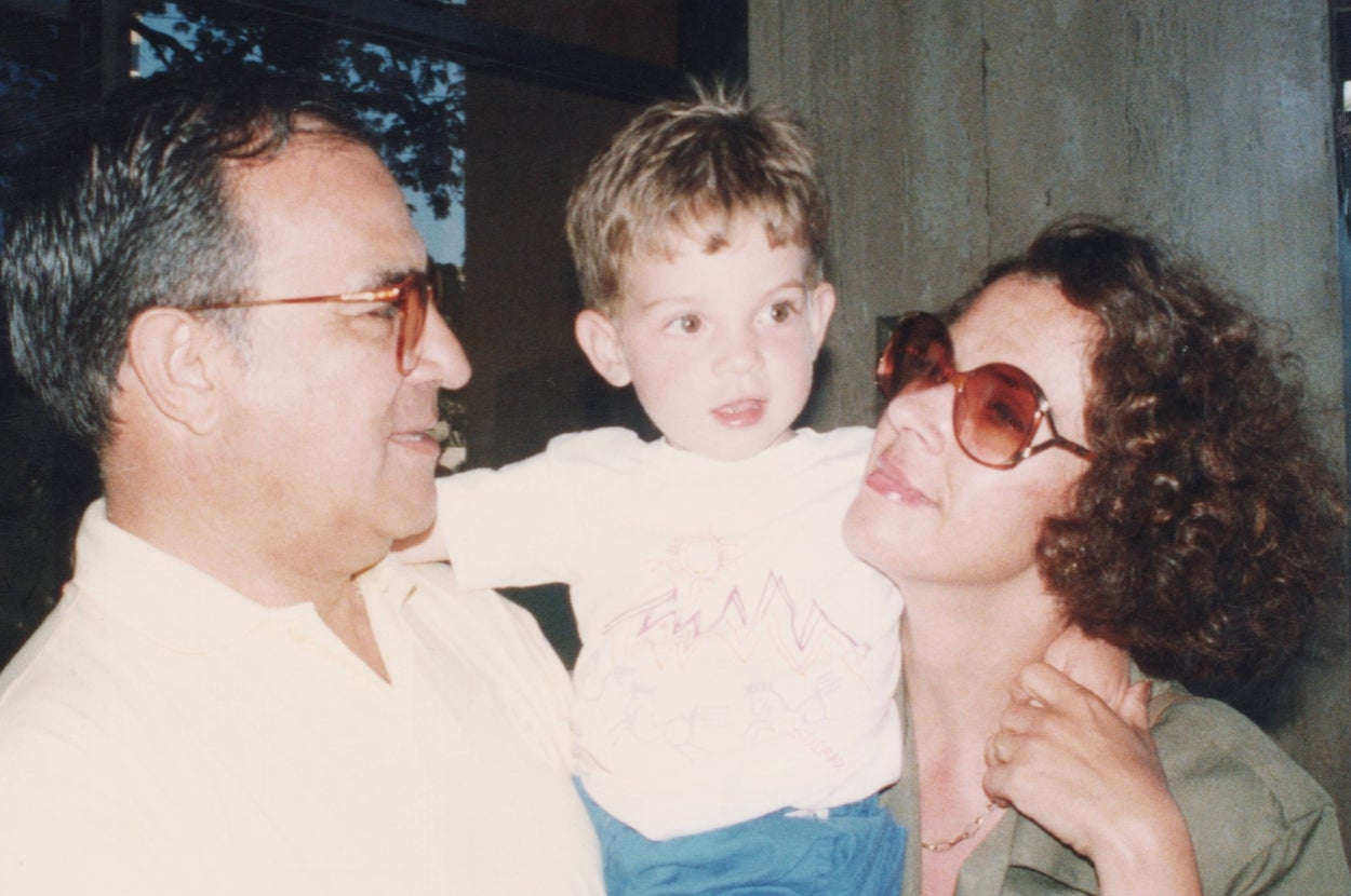An adult holding a young child with another adult standing beside them, both wearing glasses and smiling affectionately