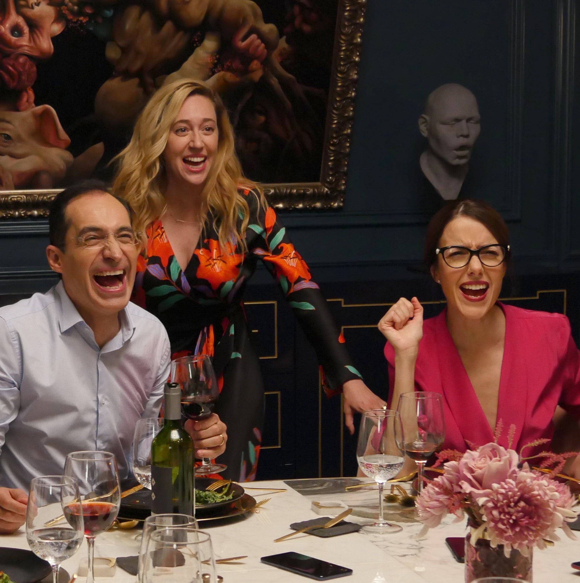 Three radical   laughing astir   a eating  array  with vino  glasses. A pistillate   successful  a floral formal  and different  successful  a pinkish  apical  are visible. A coating  is successful  the background
