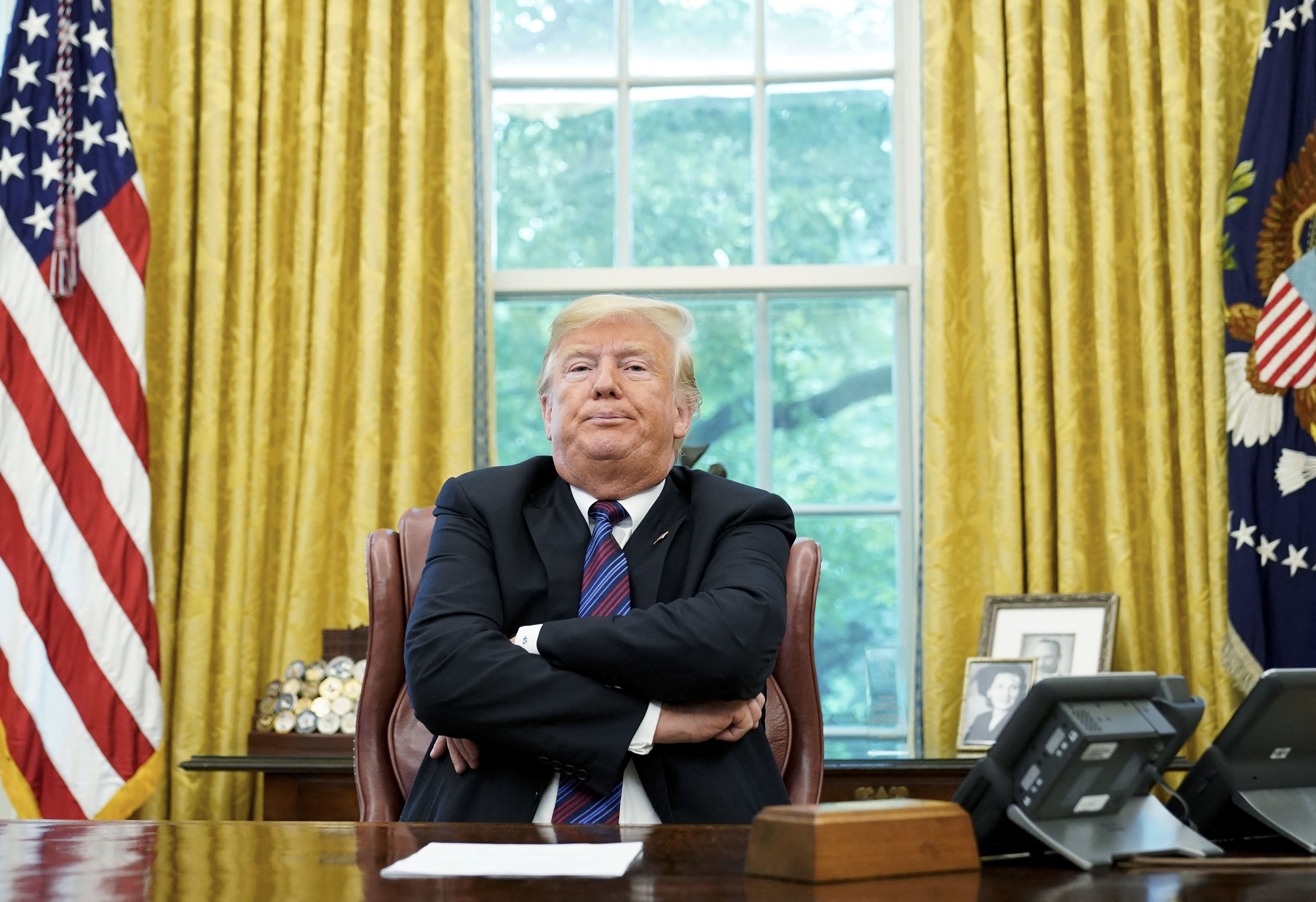 Orang yang duduk di meja kantor resmi, lengan disilangkan, diapit oleh bendera Amerika dan stempel presiden