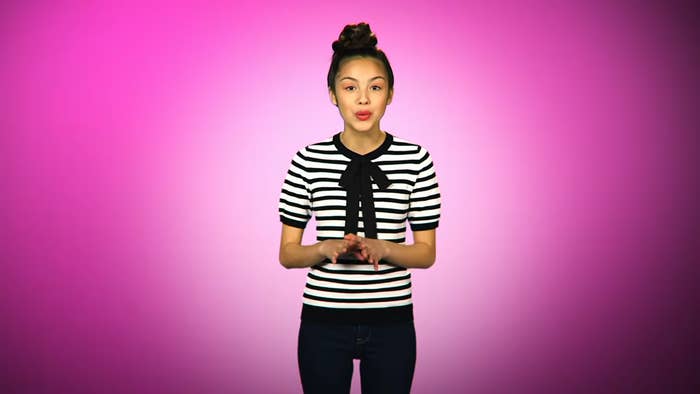 Person standing against a pink background, wearing a black and white striped shirt with a bow detail, and dark pants