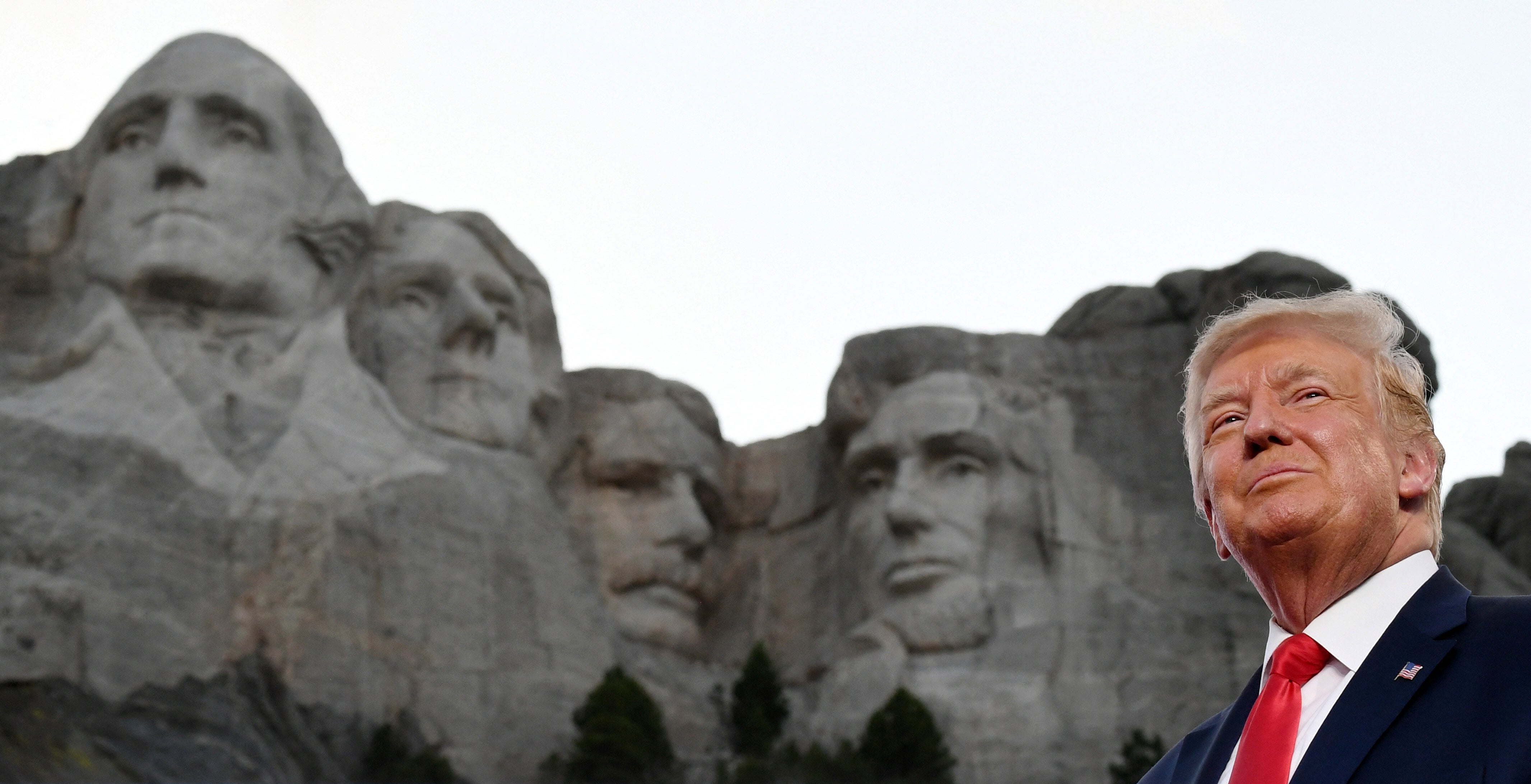 Seseorang berdiri di depan Gunung Rushmore, mengenakan jas, menatap ke atas sambil tersenyum tipis