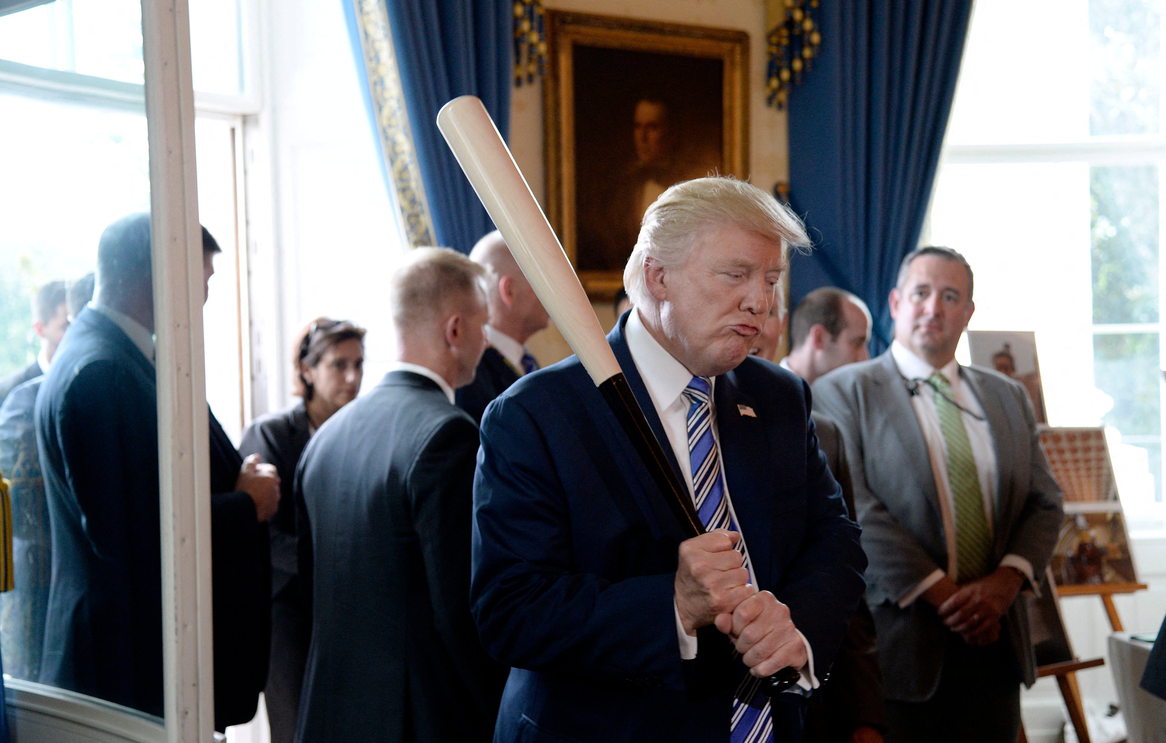 Seorang pria berjas memegang tongkat baseball di ruangan formal dengan orang-orang di latar belakang
