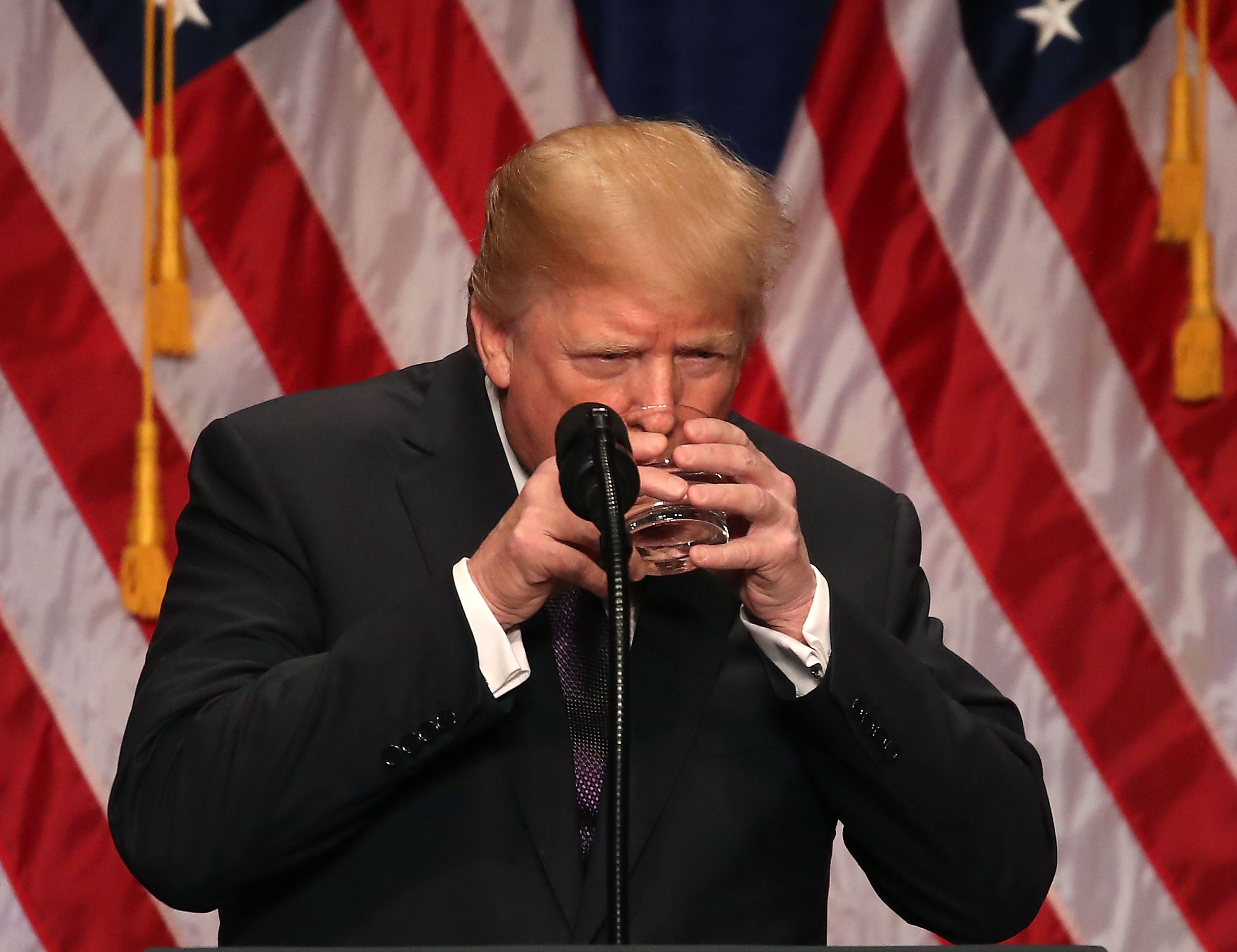 Orang berjas meminum air menggunakan kedua tangan di podium dengan latar belakang bendera Amerika
