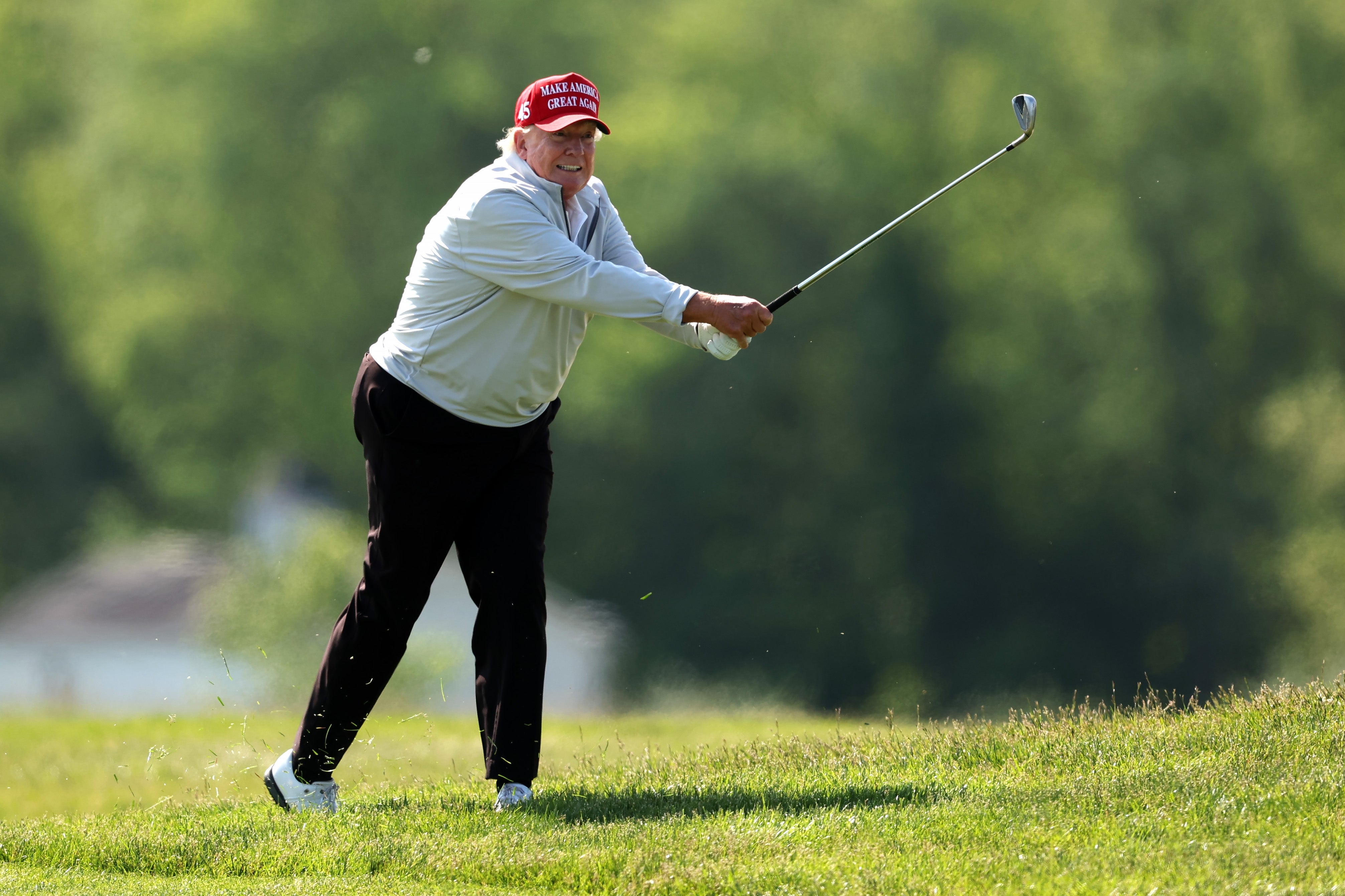 Orang yang bermain golf di lapangan berumput, mengenakan topi, kemeja lengan panjang, dan celana, melakukan ayunan tengah dengan ekspresi terfokus