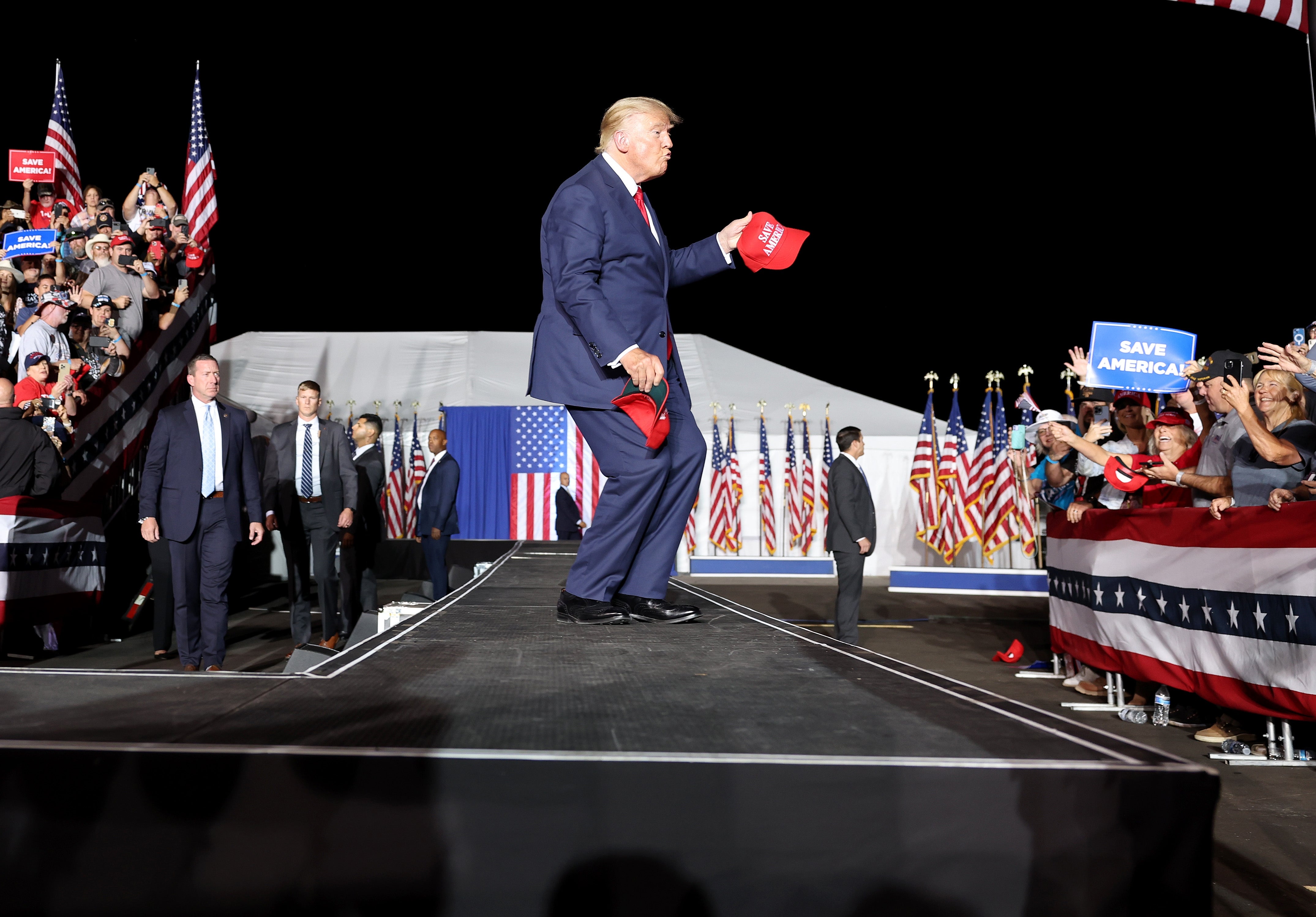 Pria berjas di atas panggung pada rapat umum memegang topi merah, dikelilingi oleh orang-orang dan bendera Amerika