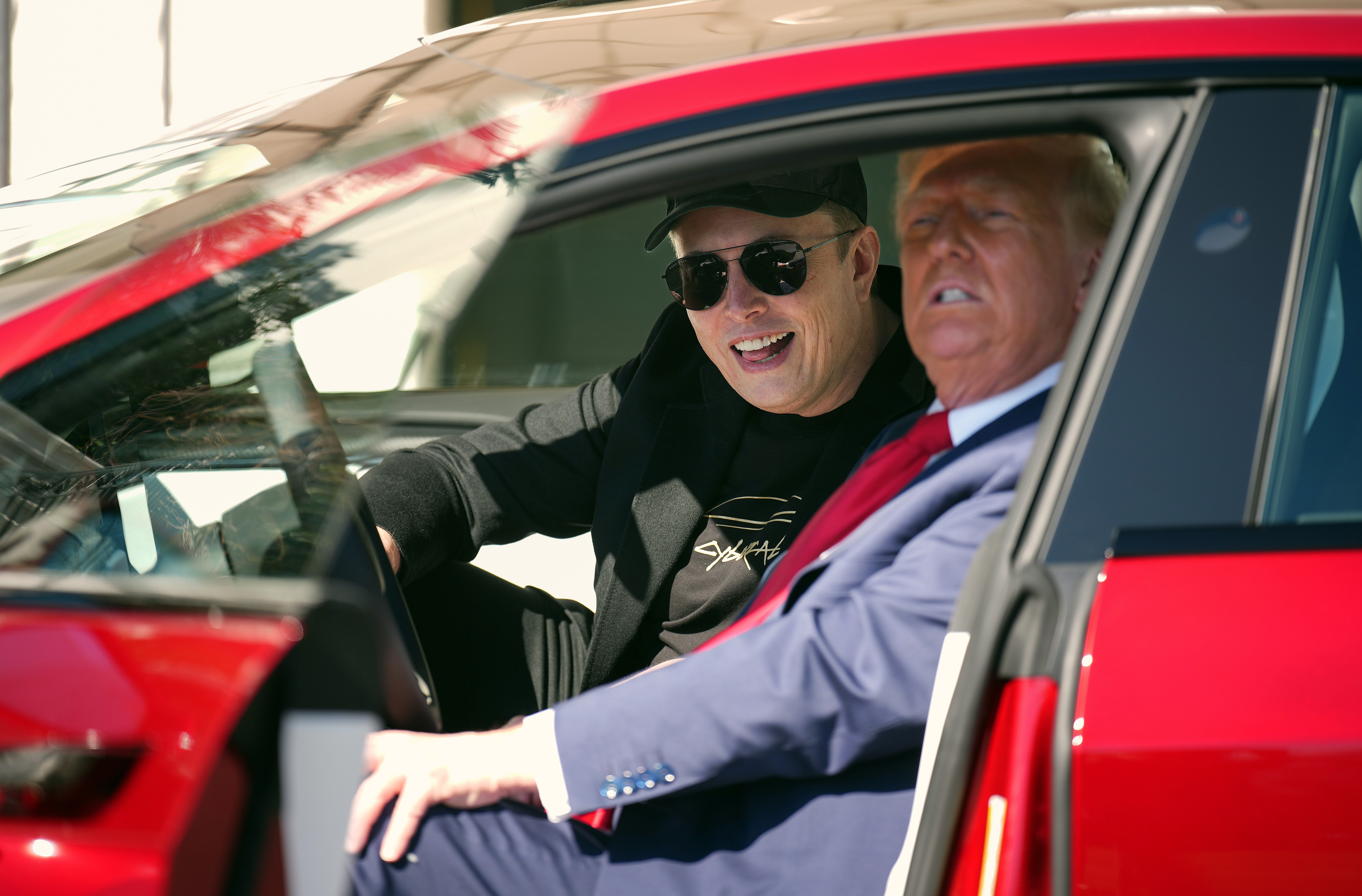 Dua laki-laki duduk di dalam mobil, yang satu memakai topi dan kacamata hitam, yang satu lagi berjas formal dan dasi merah, tersenyum dan berbicara