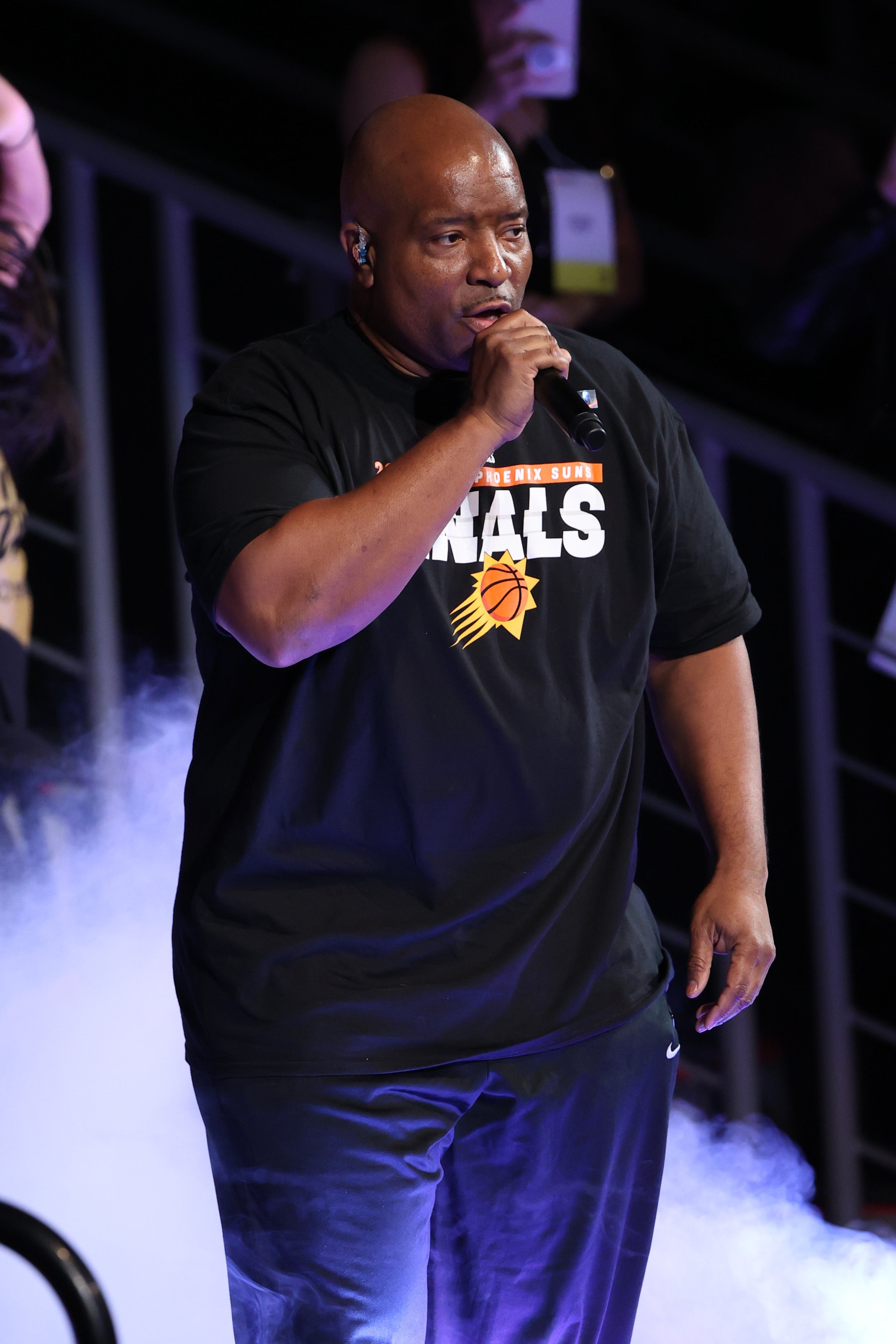 Orang di atas panggung mengenakan kemeja kasual berlogo Phoenix Suns, berbicara melalui mikrofon, dengan asap panggung di sekelilingnya