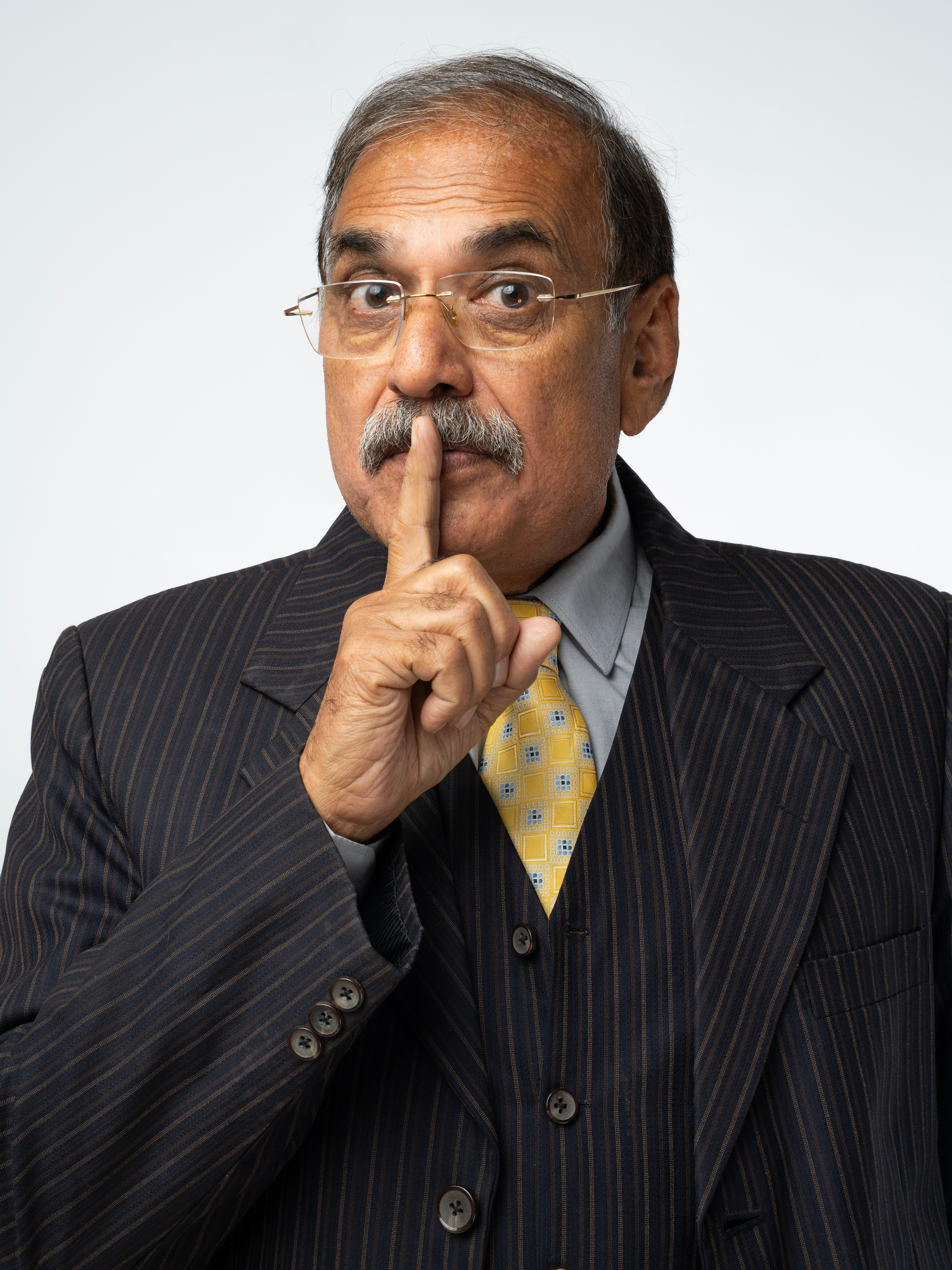 Man successful  a pinstripe suit   holds a digit  to his lips, gesturing for silence, with a focused look  and wearing glasses