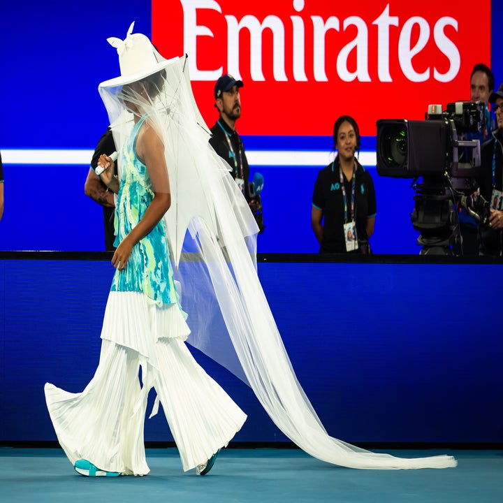 Person in a dramatic outfit with a flowing veil and pleated pants walks on a sports court, attended by staff and cameras in the background