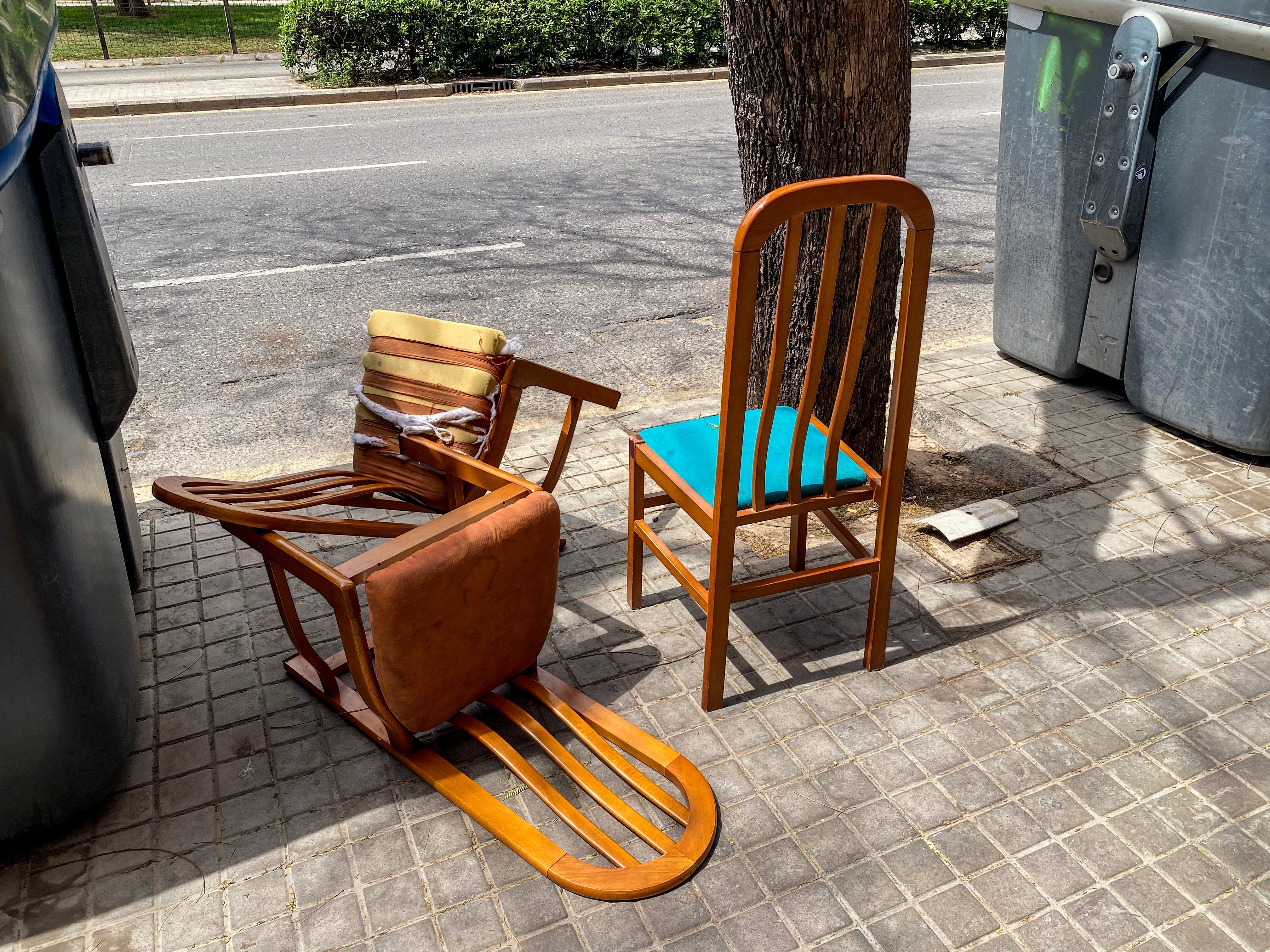Two woody  chairs connected  a sidewalk; 1  upright with a bluish  seat, the different   toppled over, adjacent   trash bins and a tree
