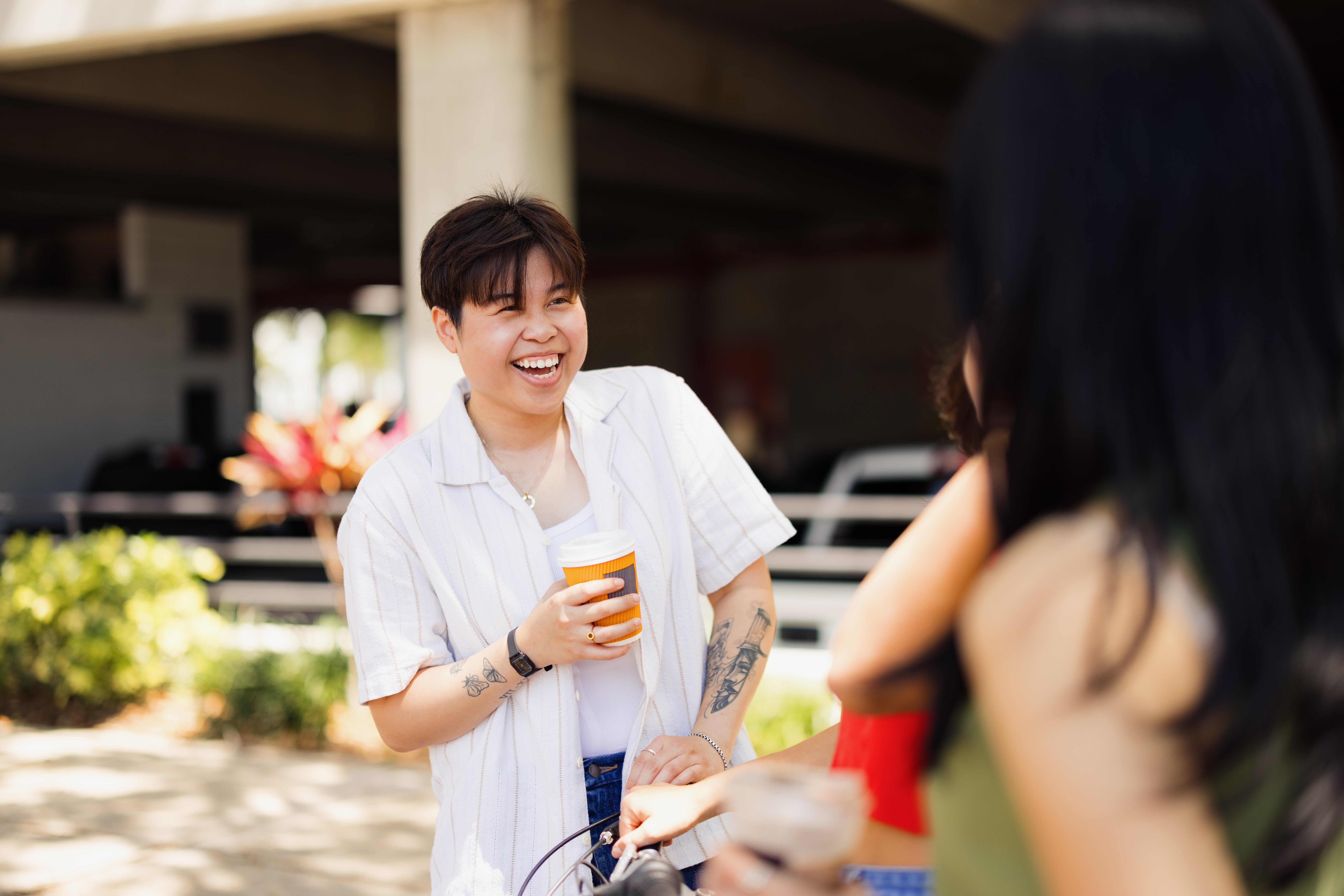 Person with abbreviated  hair, wearing a casual unfastened  shirt, smiling and holding a drink, engaged successful  speech  with friends outdoors