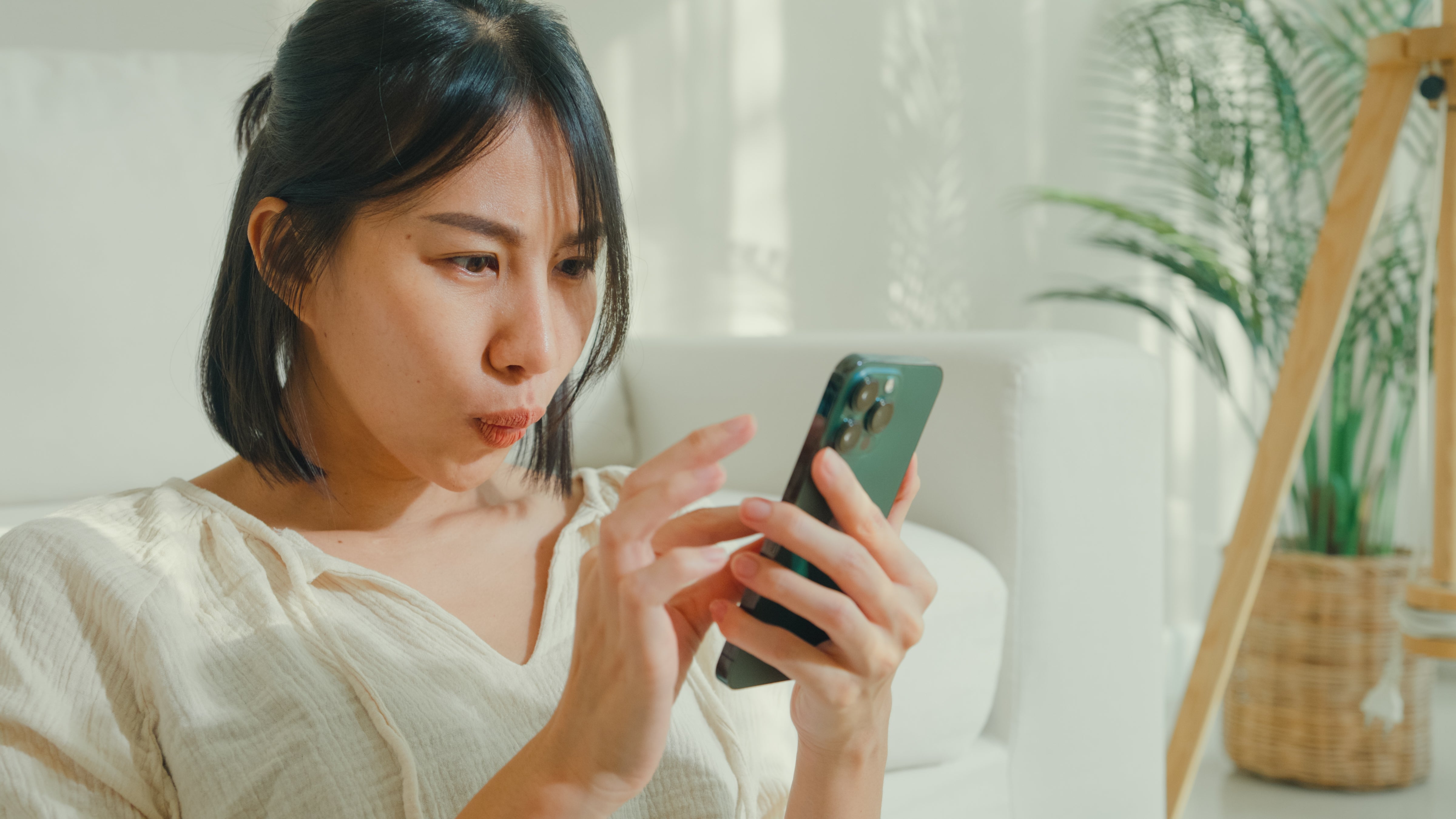 Orang yang duduk di dalam ruangan, melihat smartphone dengan penuh perhatian, dengan ekspresi terfokus. Sebuah tanaman terlihat di latar belakang
