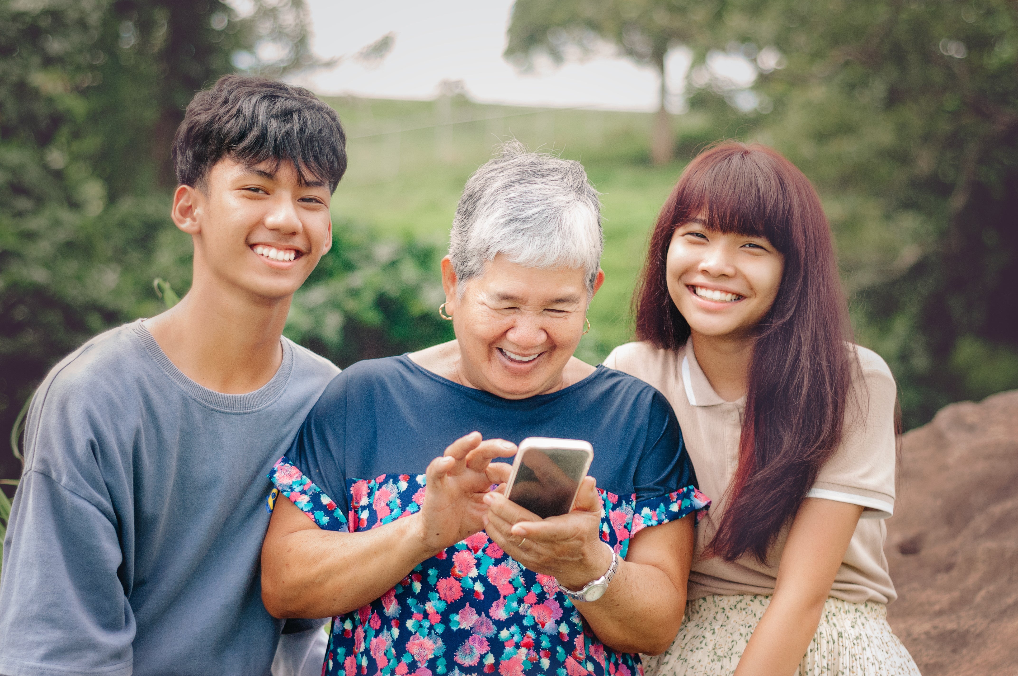 Three radical   smiling outdoors, including an aged  pistillate   looking astatine  a smartphone, flanked by a young antheral   and woman