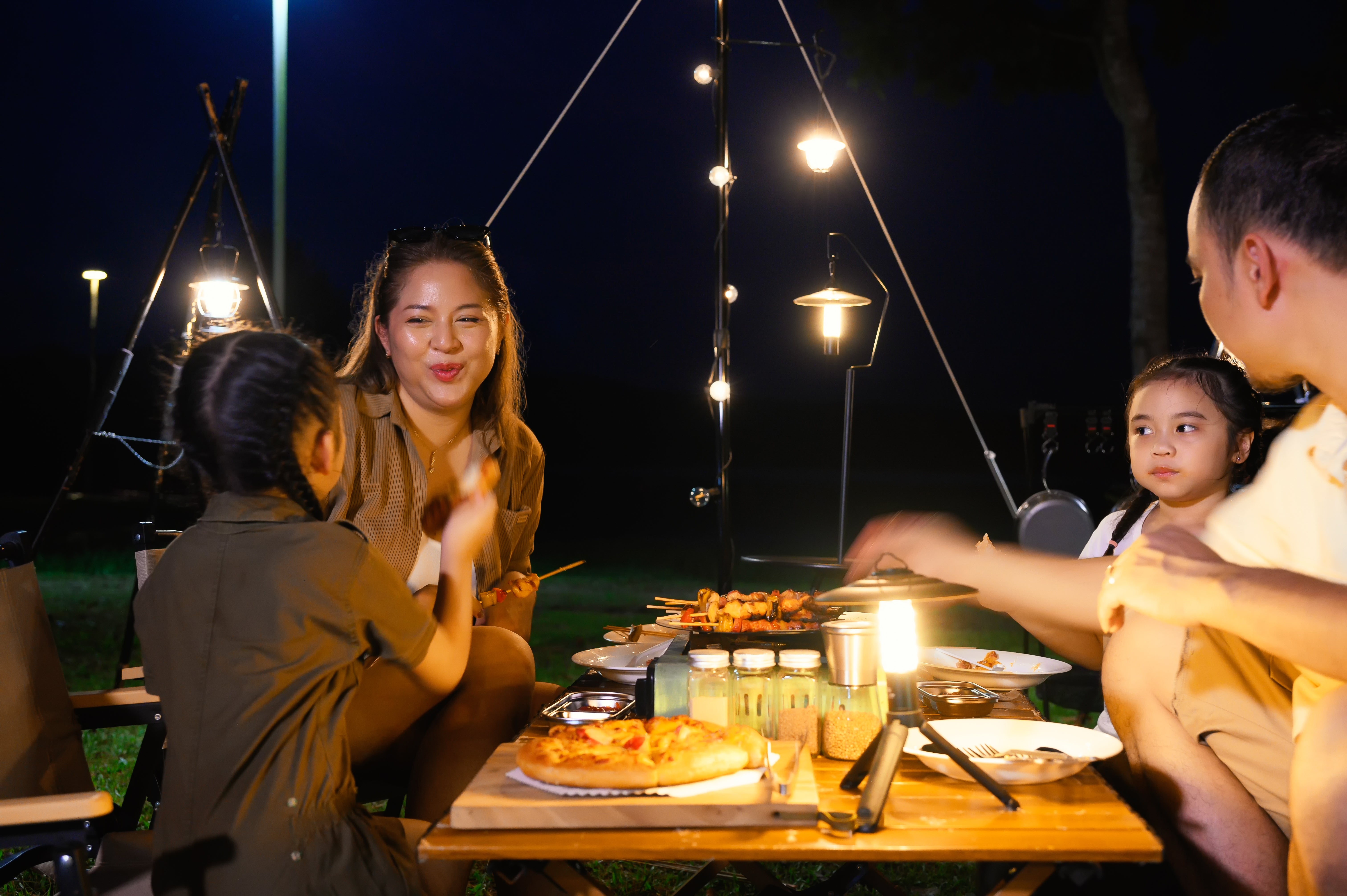 Family enjoys an outdoor nighttime  picnic with grilled nutrient  and pizza, seated astir   a array  nether  lukewarm  drawstring  lights