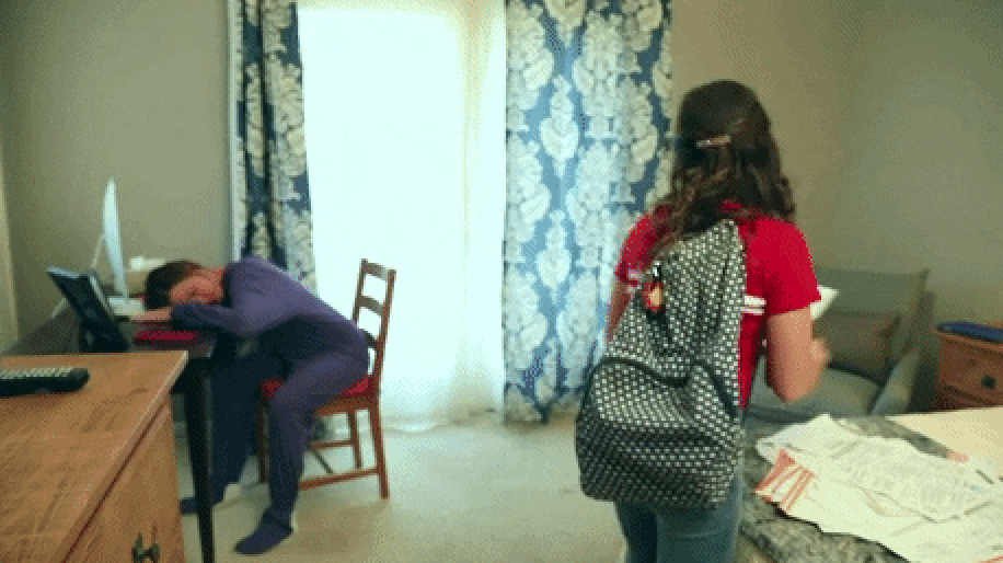 A person in a purple bodysuit sleeps at a desk; another wearing a polka dot backpack stands nearby in a bedroom