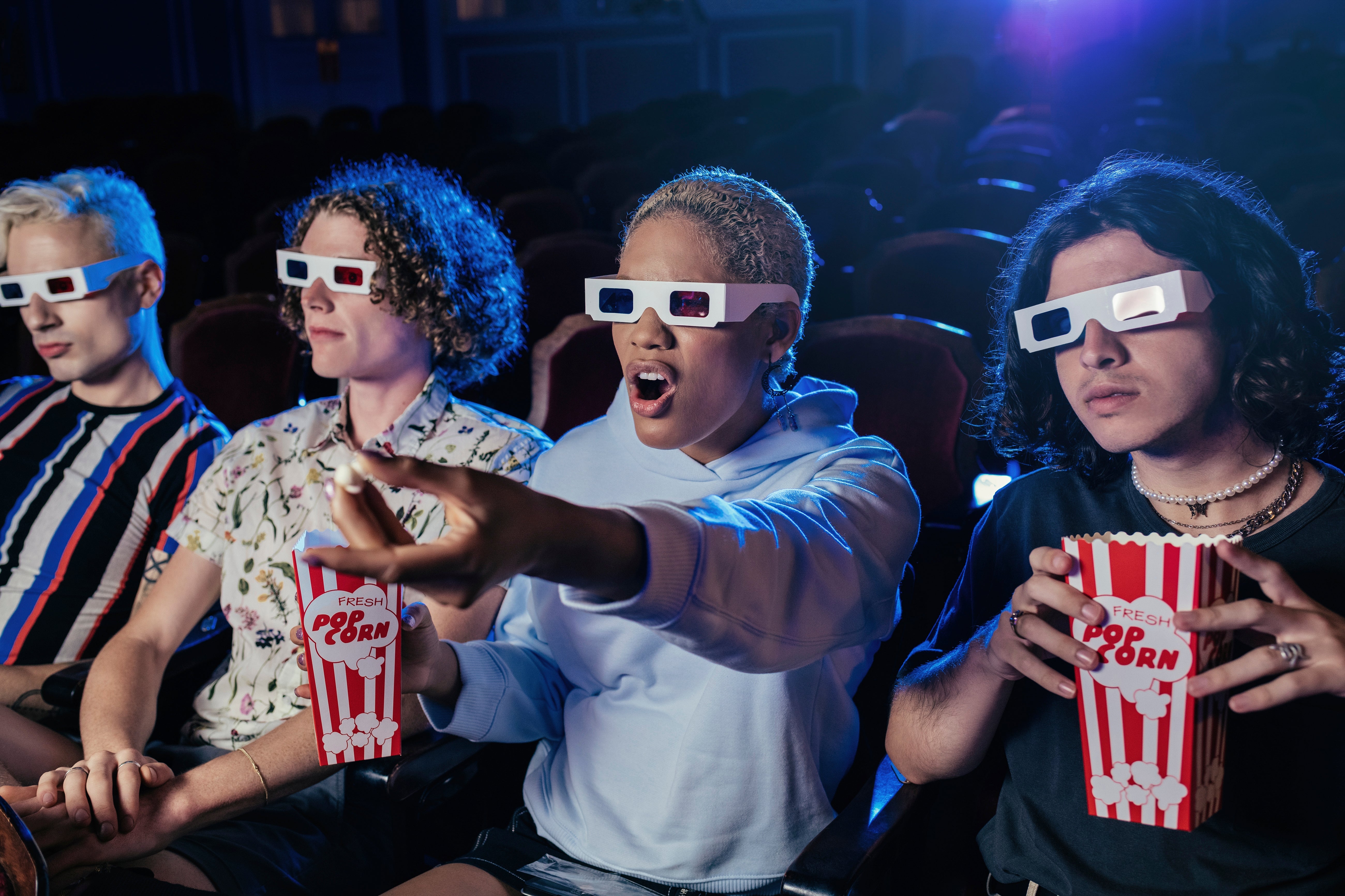 Empat orang memakai kacamata 3D, duduk di teater, memegang popcorn, dan bereaksi dengan terkejut