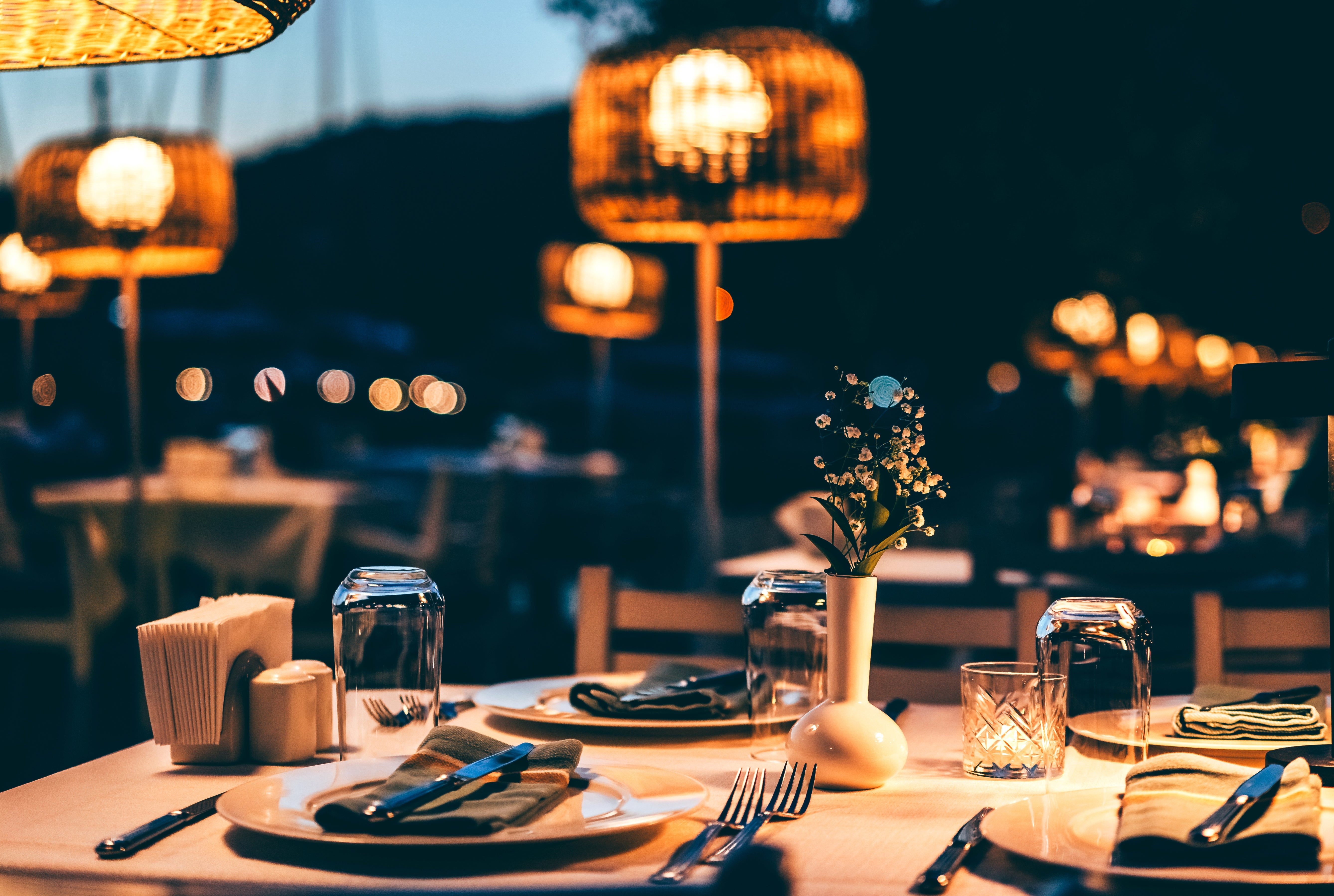 Penataan ruang makan outdoor yang elegan dengan piring, peralatan makan, dan gelas di atas meja, diterangi lampu gantung dekoratif, menciptakan suasana nyaman