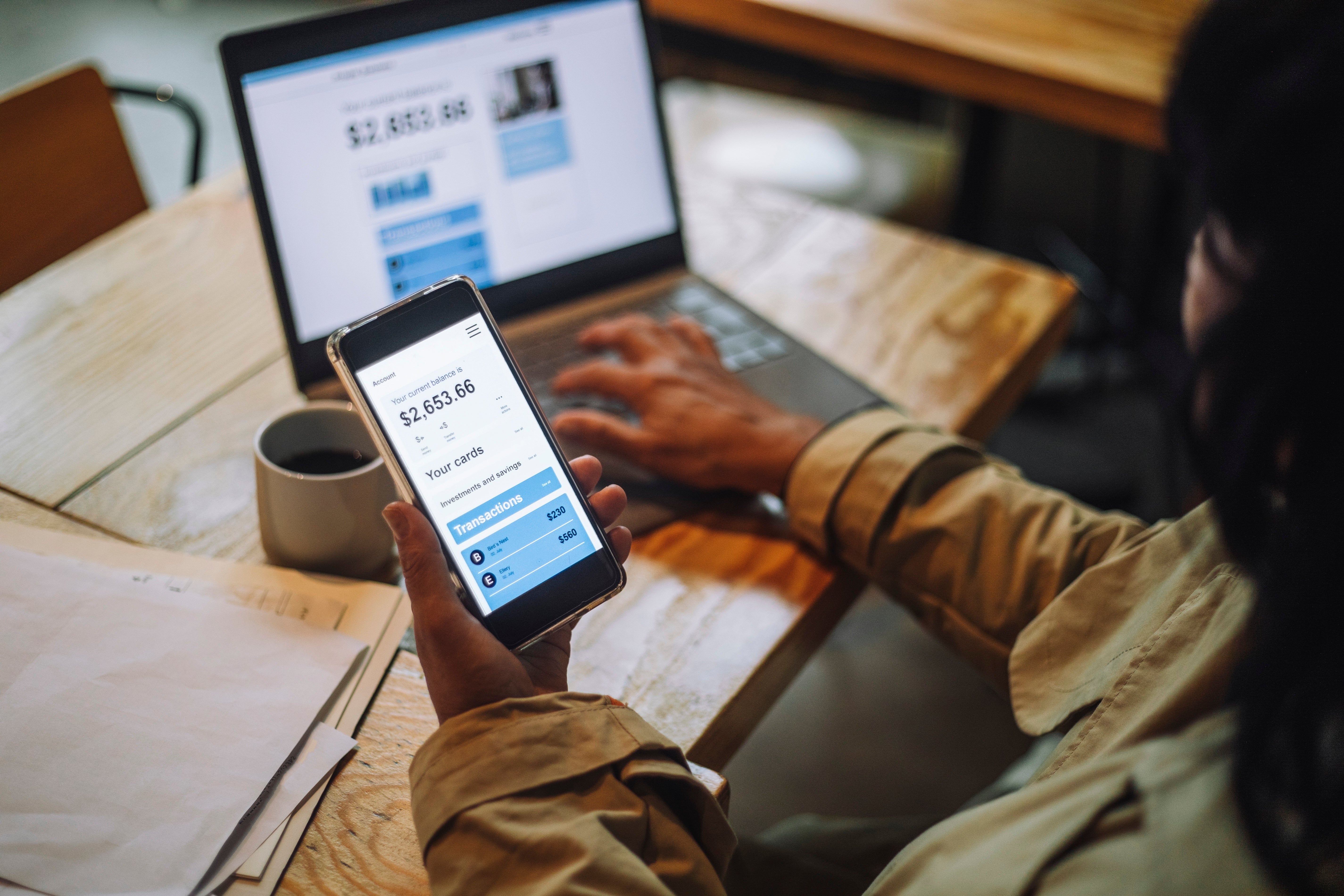 Seseorang menggunakan smartphone untuk memeriksa saldo rekening bank, dengan laptop dan cangkir kopi di atas meja kayu