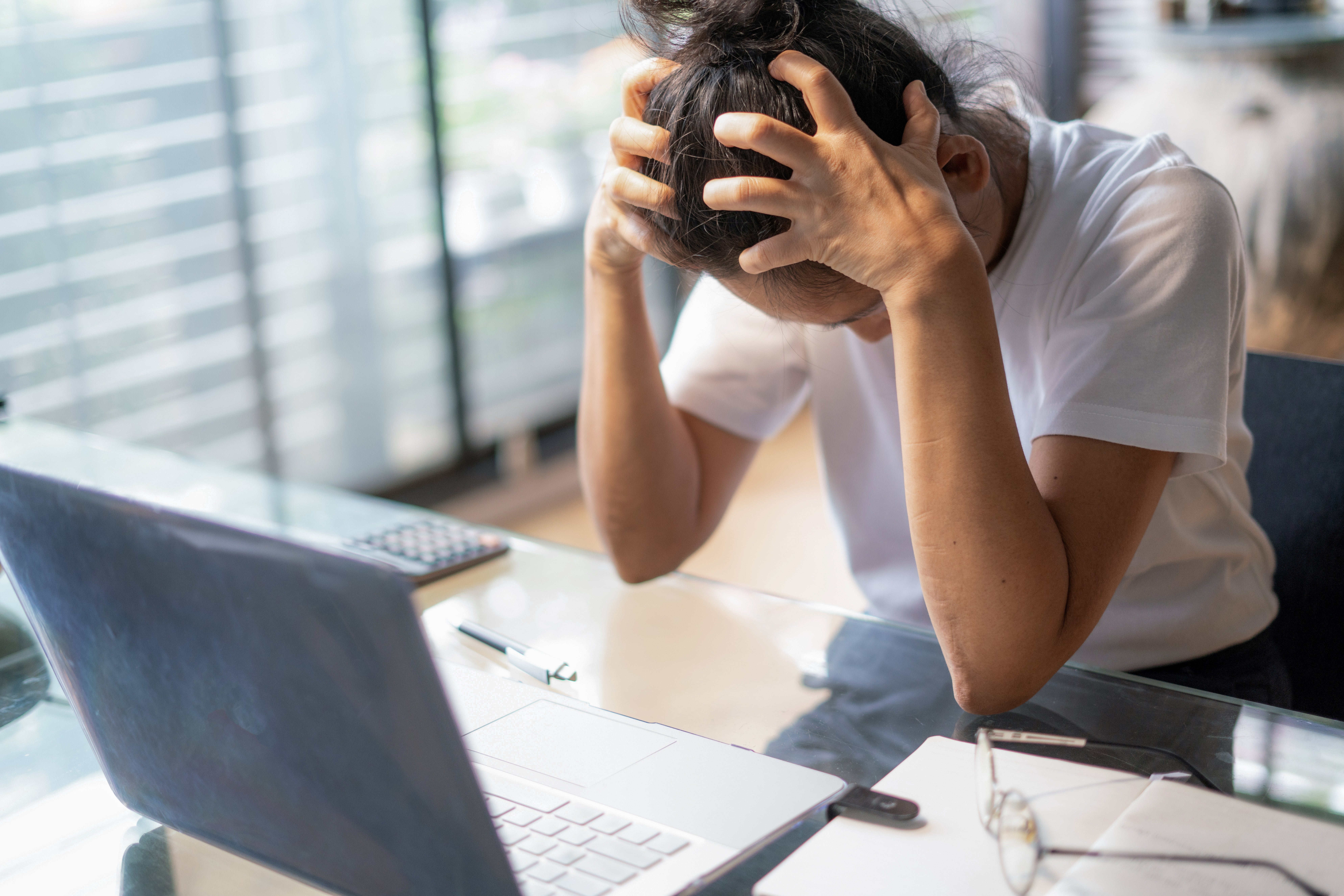 Orang yang duduk di meja dengan tangan di atas kepala karena frustrasi, melihat laptop