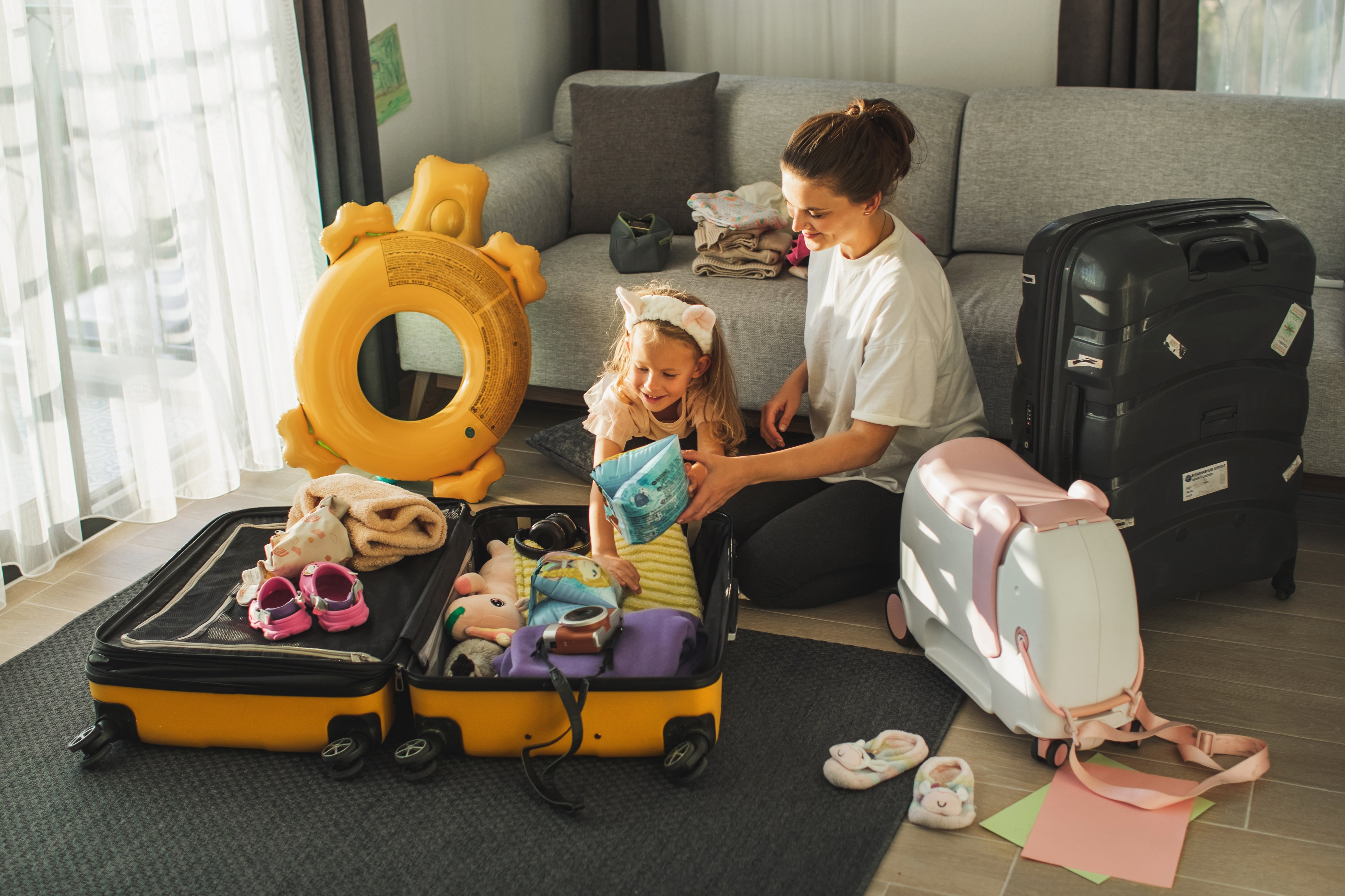 A woman and child pack a suitcase in a living room. A suitcase, inflatable float, and toys are nearby