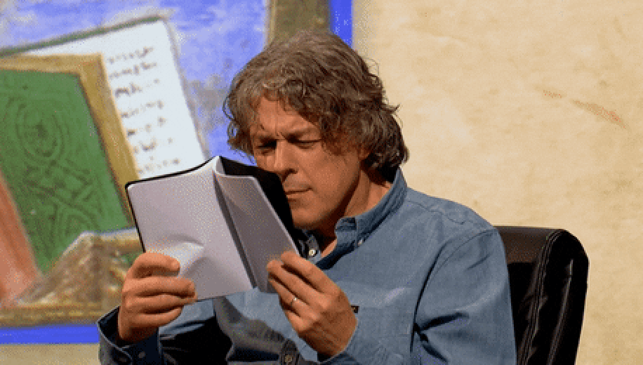 Person in a denim shirt squinting at papers, seated with a book and abstract art in the background