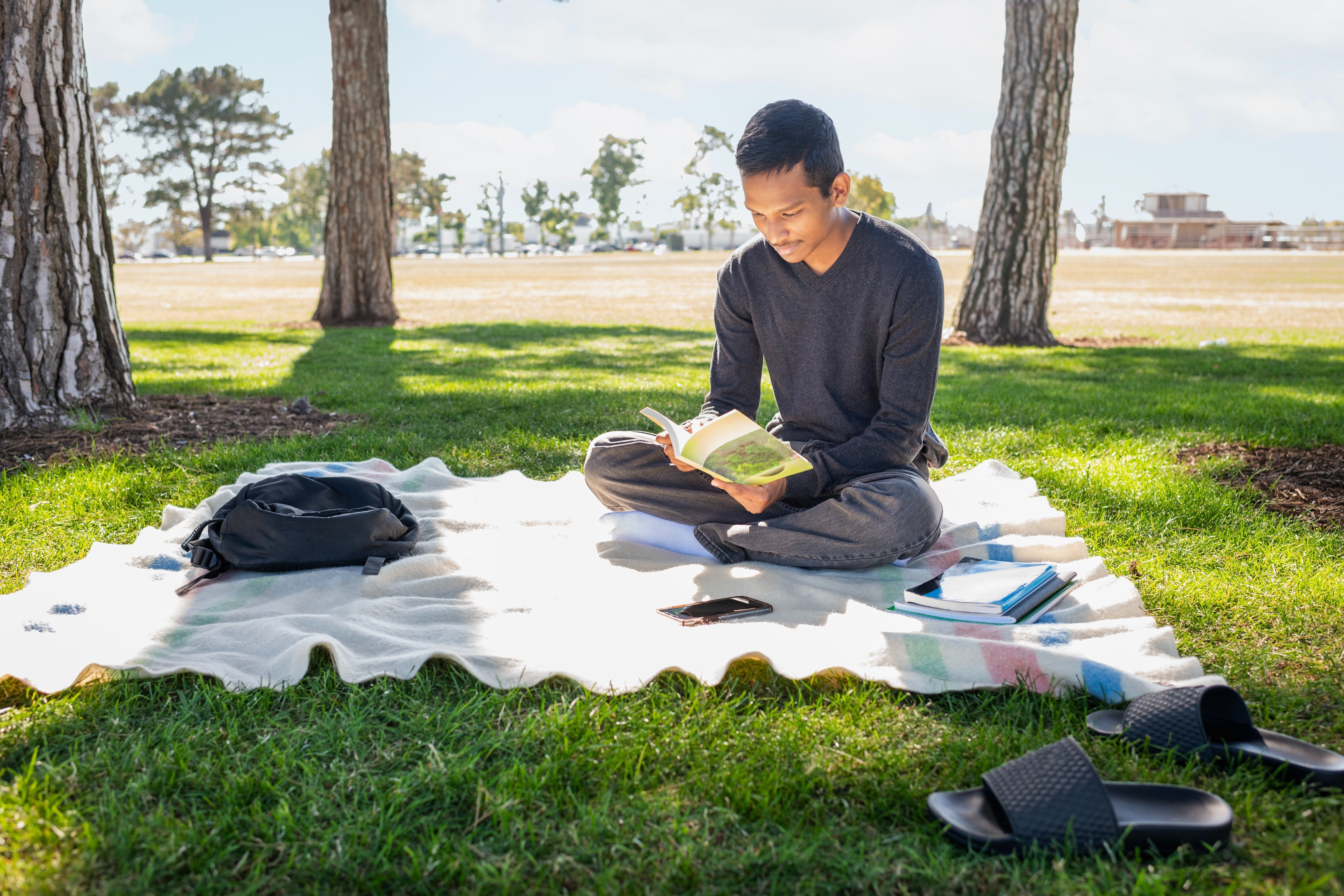 Person sitting cross-legged on a blanket in a park, reading a book, with a backpack and sandals nearby
