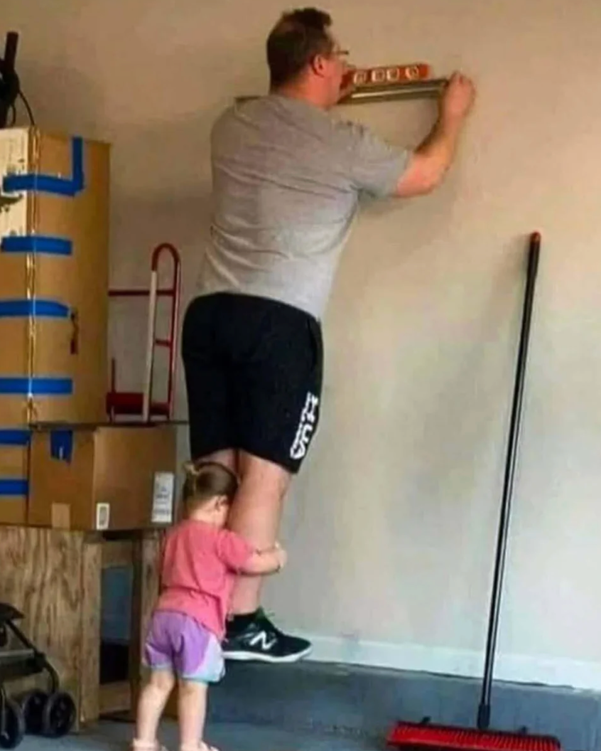 A man uses a level on a wall while a child playfully holds his leg. The scene is in a casual home setting