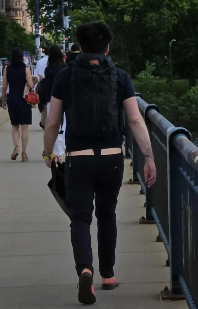 Person walking on a sidewalk, wearing a backpack and carrying a bag, with a humorous optical illusion showing a skin-tone waistband resembling bare skin