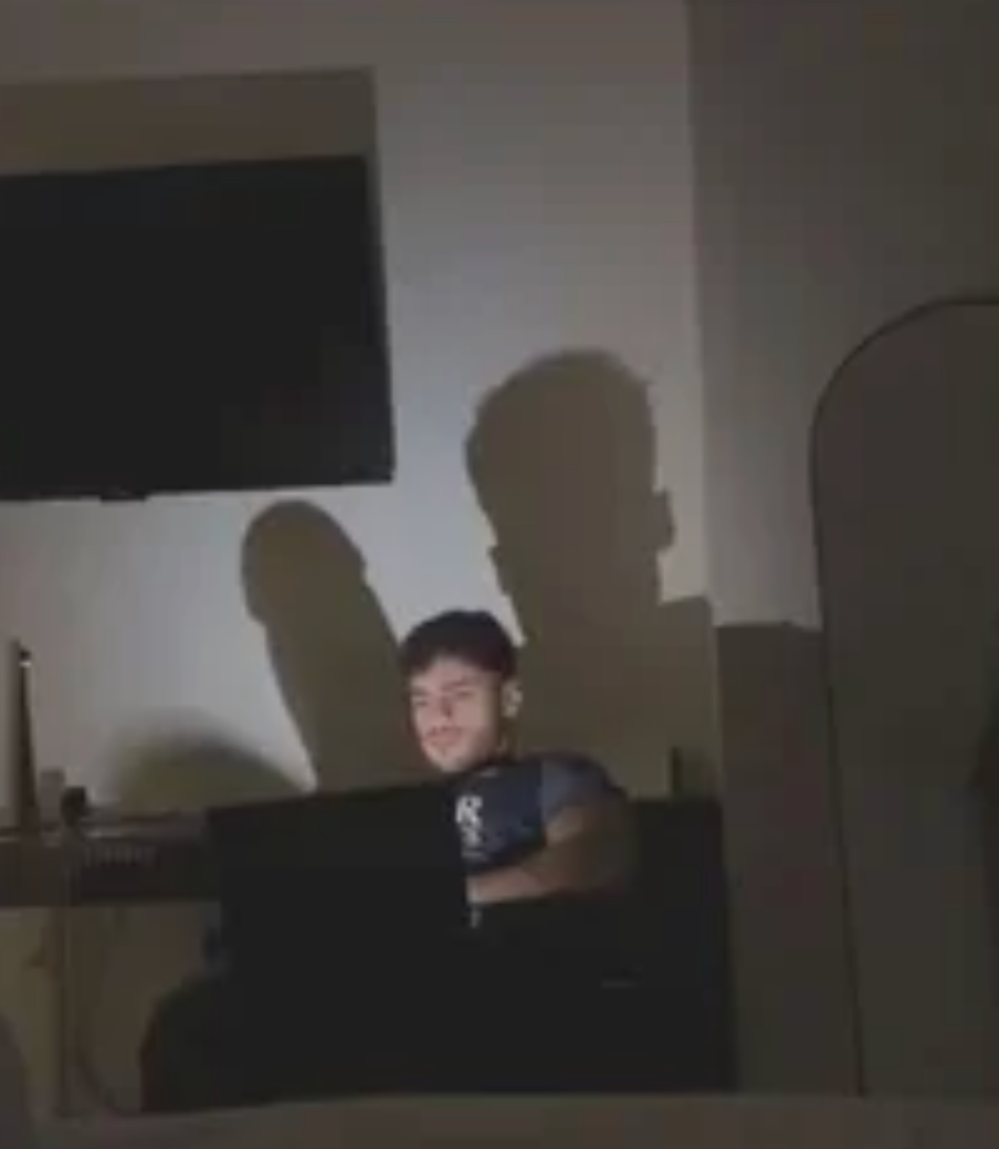 Person sitting in a dimly lit room, using a laptop. A shadow on the wall is humorously shaped like a dinosaur
