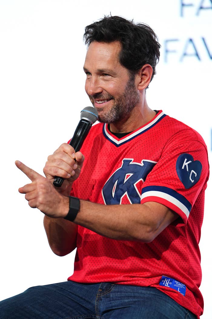 Paul Rudd in a jersey with "KC" on the sleeve speaking into a microphone on stage, gesturing with a smile