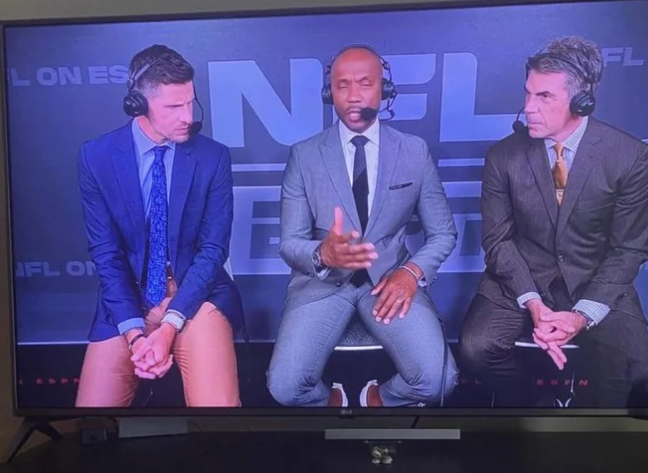 Three sports commentators in suits discuss on an ESPN set. The man in the middle gestures while speaking