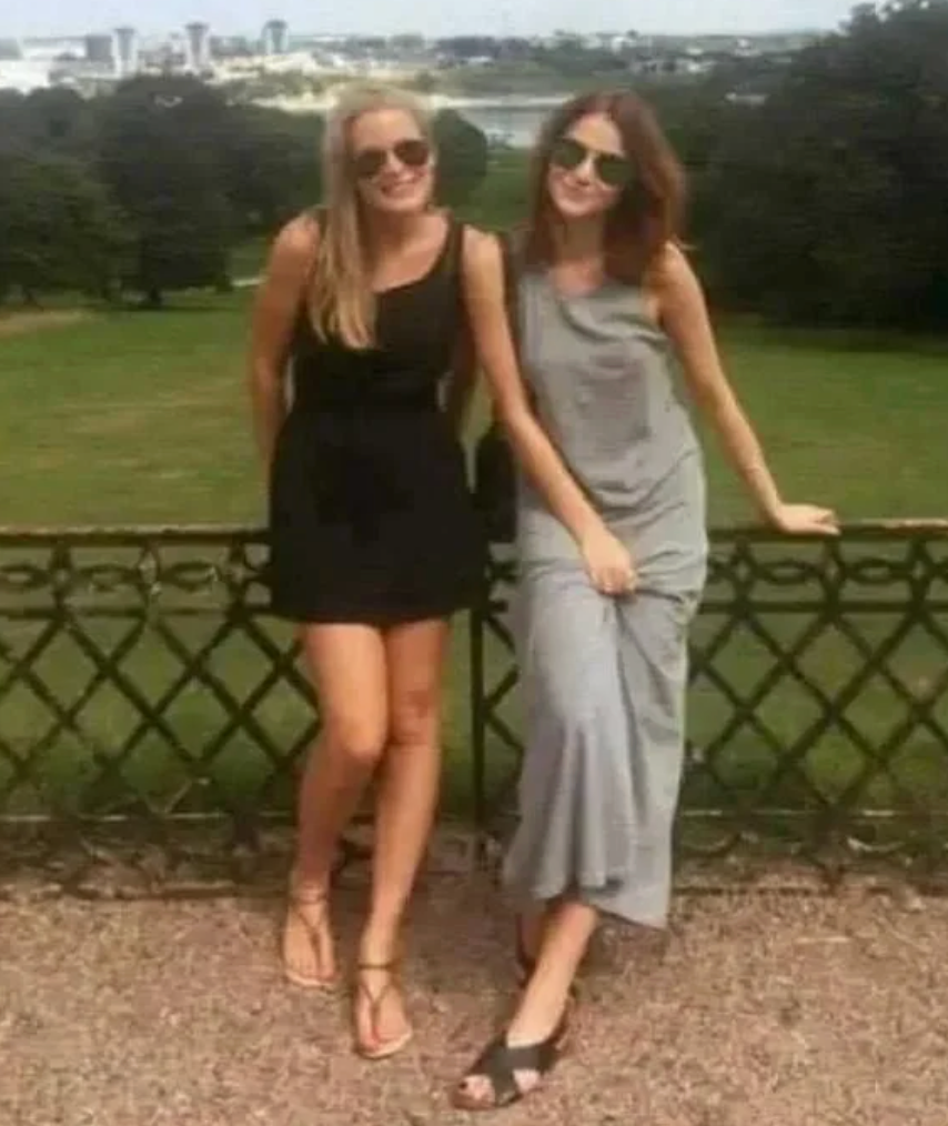 Two women in sunglasses and casual dresses stand by a fence in a park with a cityscape in the background
