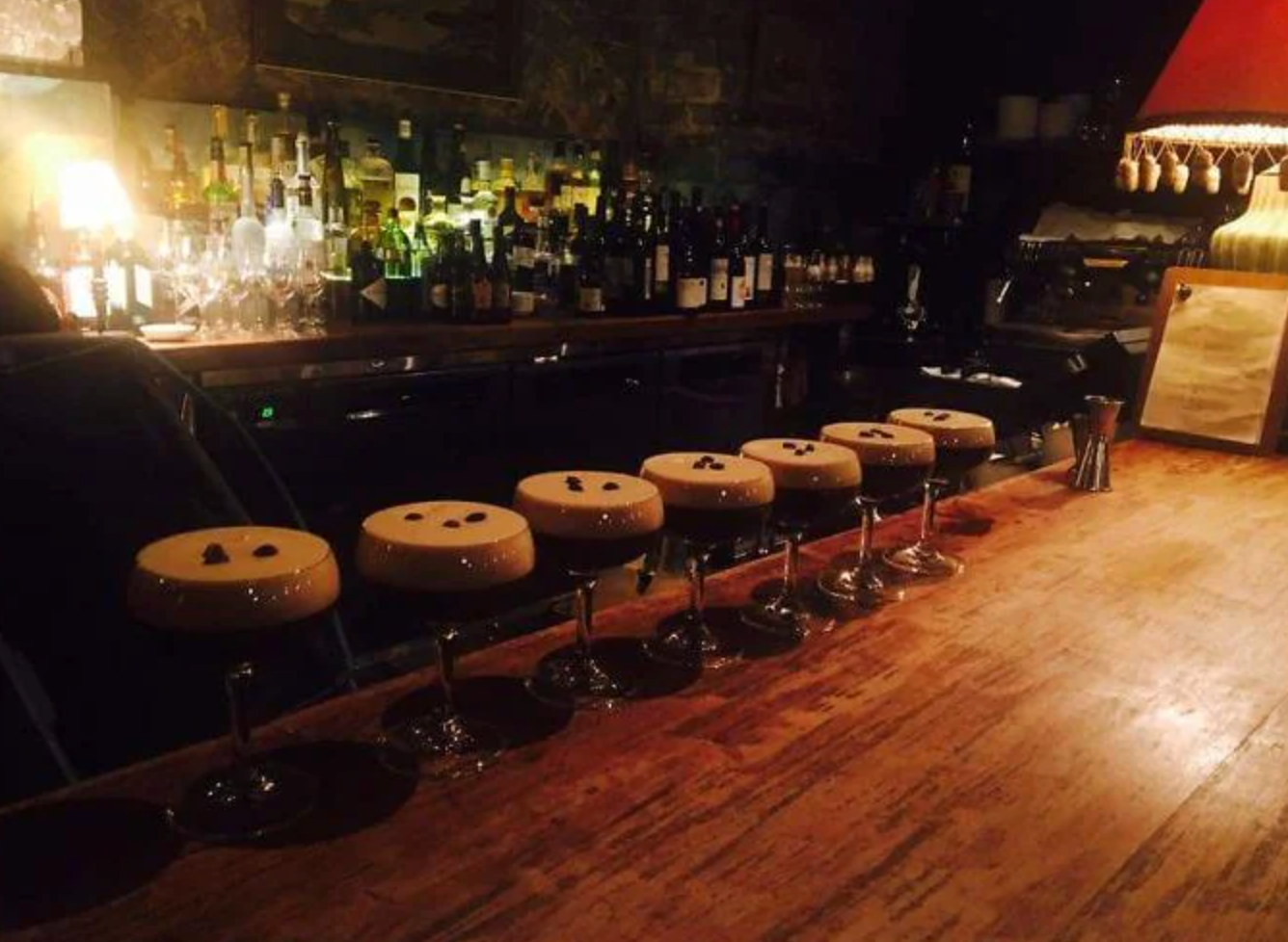Row of espresso martinis on a wooden bar counter with bottles and dim lighting in the background