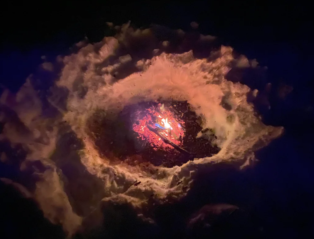 A spiral-shaped snow pit with a small fire burning at its center, creating a warm glow amidst the surrounding snow
